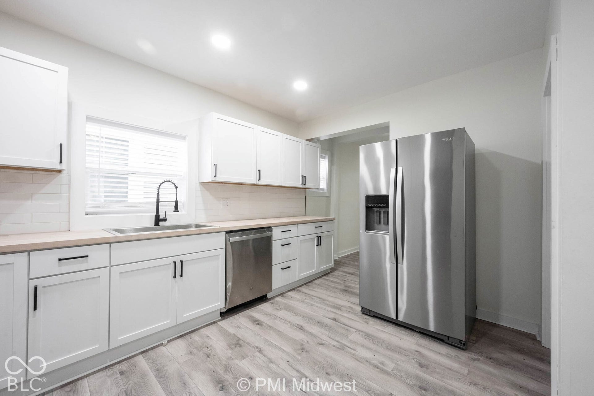 White shaker cabinetry with matte black hardware, stainless steel appliances, stainless steel sink with a black gooseneck faucet, light wood-finish countertops, and wood-finish flooring