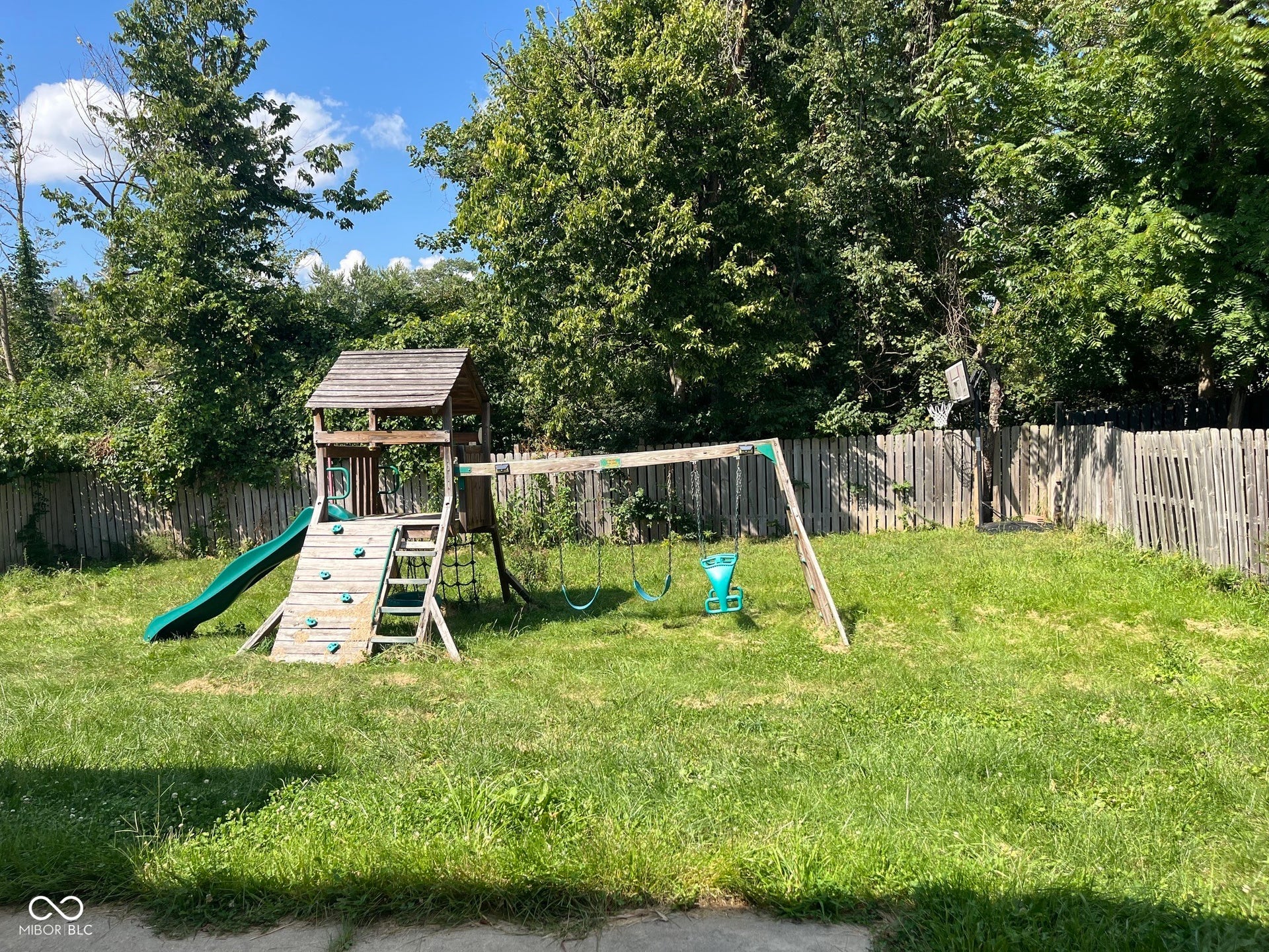 Expansive fenced backyard featuring a wooden playset with a slide and swings, surrounded by mature trees