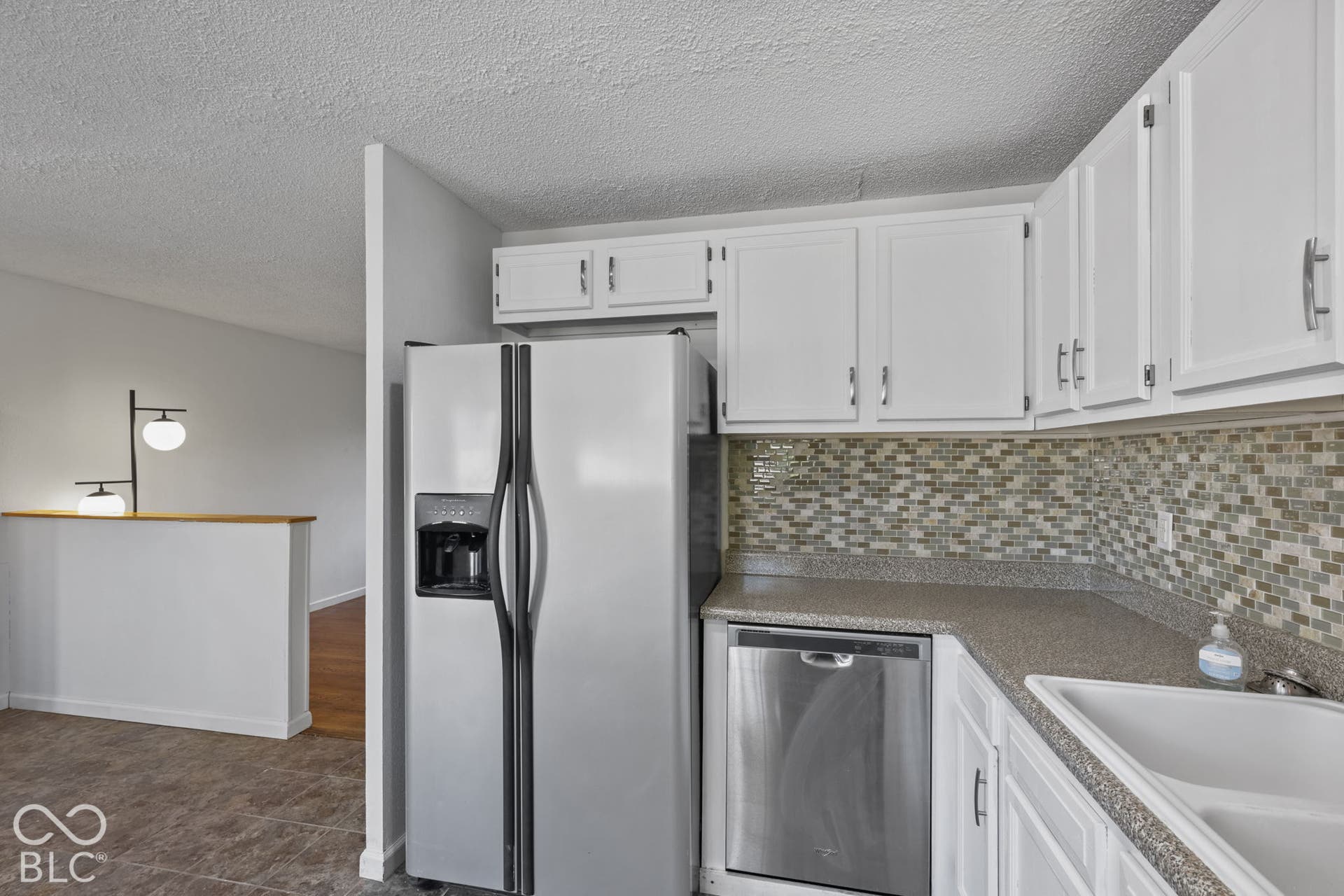 White cabinetry with brushed nickel hardware, mosaic tile backsplash, solid surface countertops, stainless steel dishwasher, and a stainless steel refrigerator