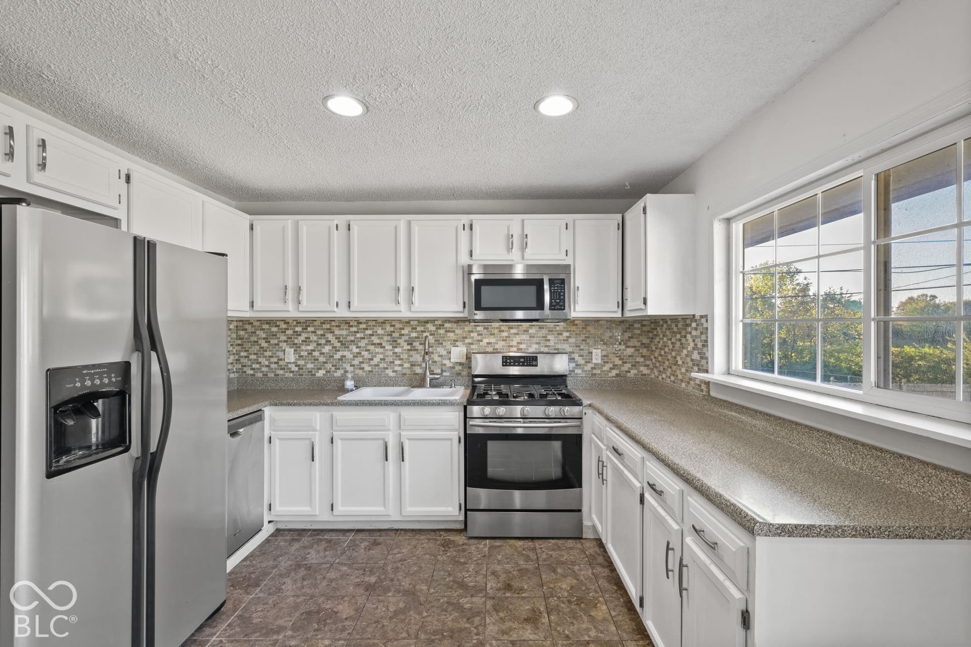White cabinetry, stainless steel appliances, patterned backsplash, light-colored countertops, and a large window