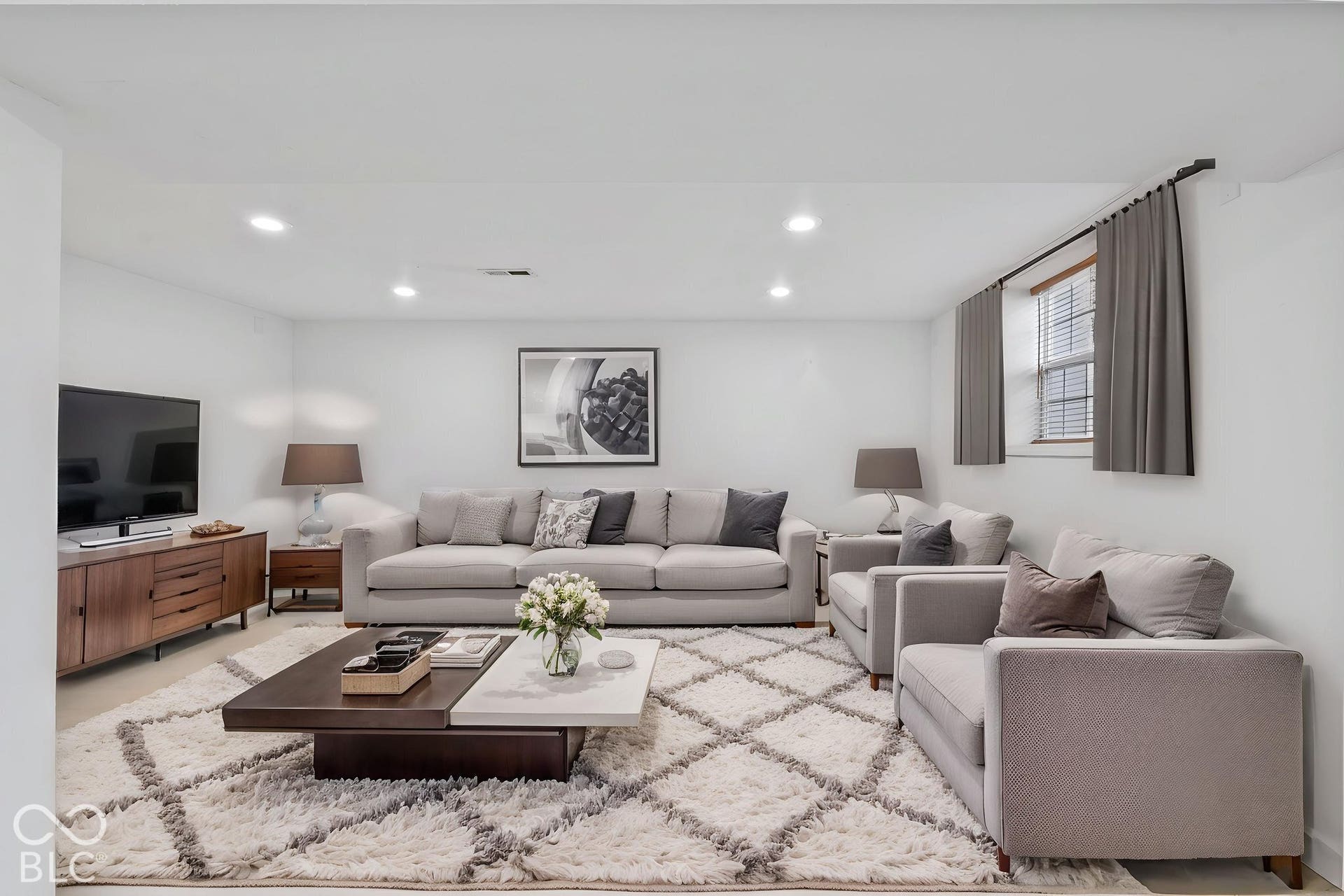 Recessed ceiling lighting illuminates a spacious living area featuring a single window with blinds, a built-in media console, and light-toned walls