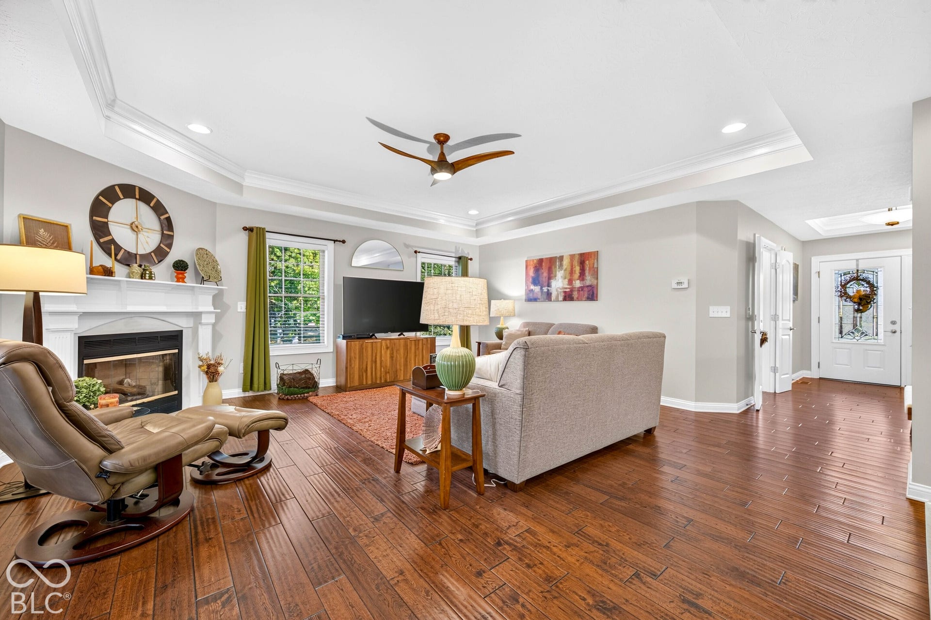 Hardwood-finish flooring throughout, tray ceiling with recessed lighting, white fireplace with mantle, ceiling fan, and white front door with decorative glass inserts
