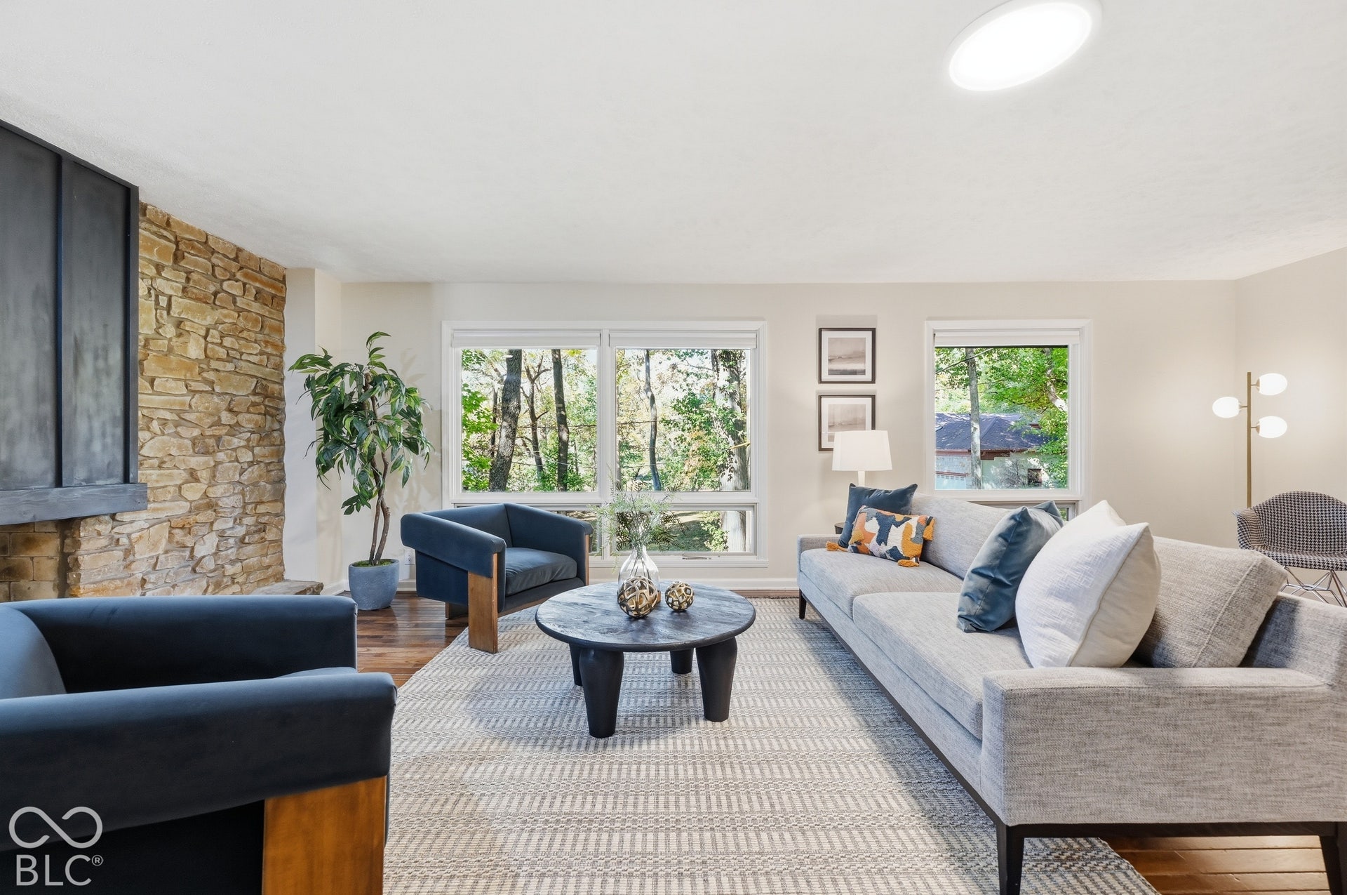 Stacked stone meets dark wood at the fireplace, a material palette from the organic modern tradition of Bruce Goff and E. Fay Jones, with the woods framed in the windows beyond.