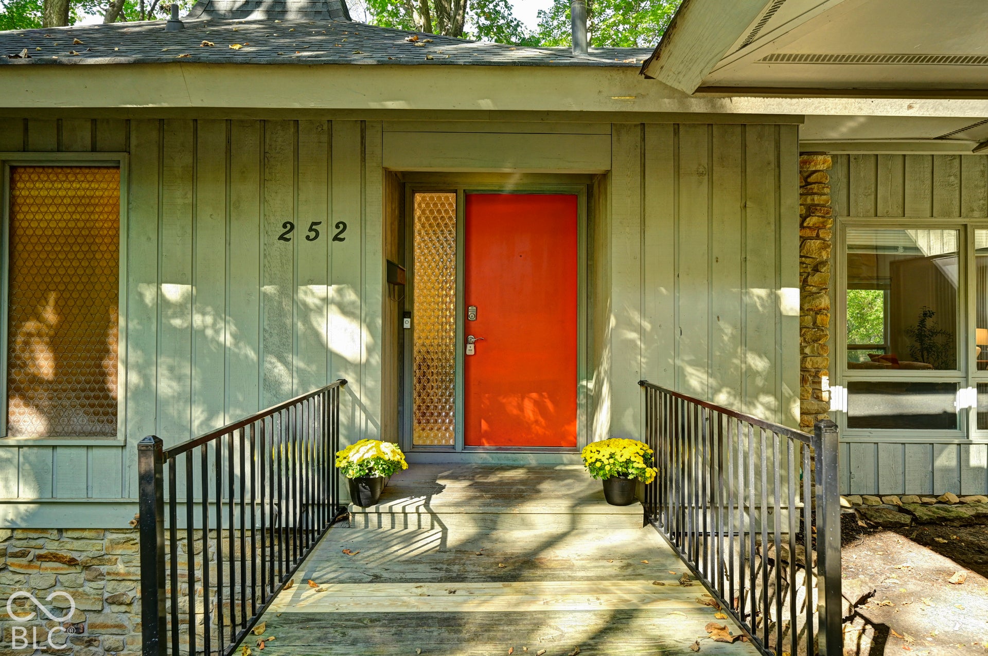 Arrival: a footbridge leads to the bold orange front door, framed by vertical wood siding and stone. The house sits into the land, just as Clyde E. Woods intended.