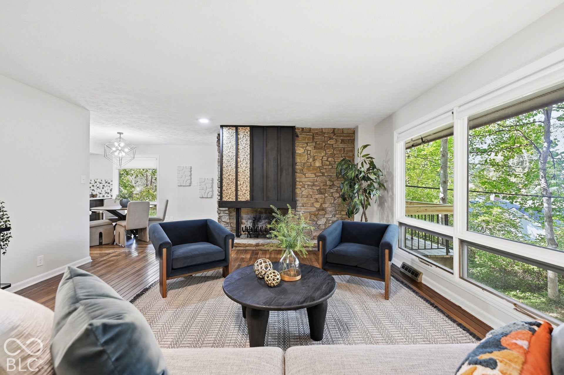 Living room to dining room in one continuous space wrapped in glass and trees, with the two-sided stone and wood fireplace defining the rooms while keeping them open. Organic modern design in Indianapolis.