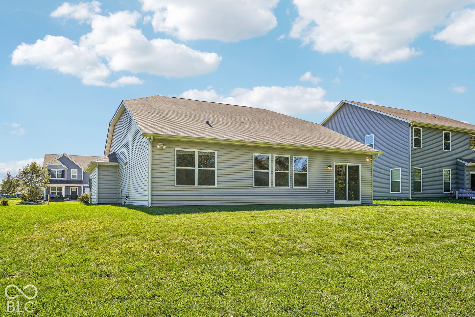 Spacious rear elevation featuring vinyl siding in soft green tones, multiple large windows with dark frames, an arched roofline, and expansive manicured lawn with mature landscaping