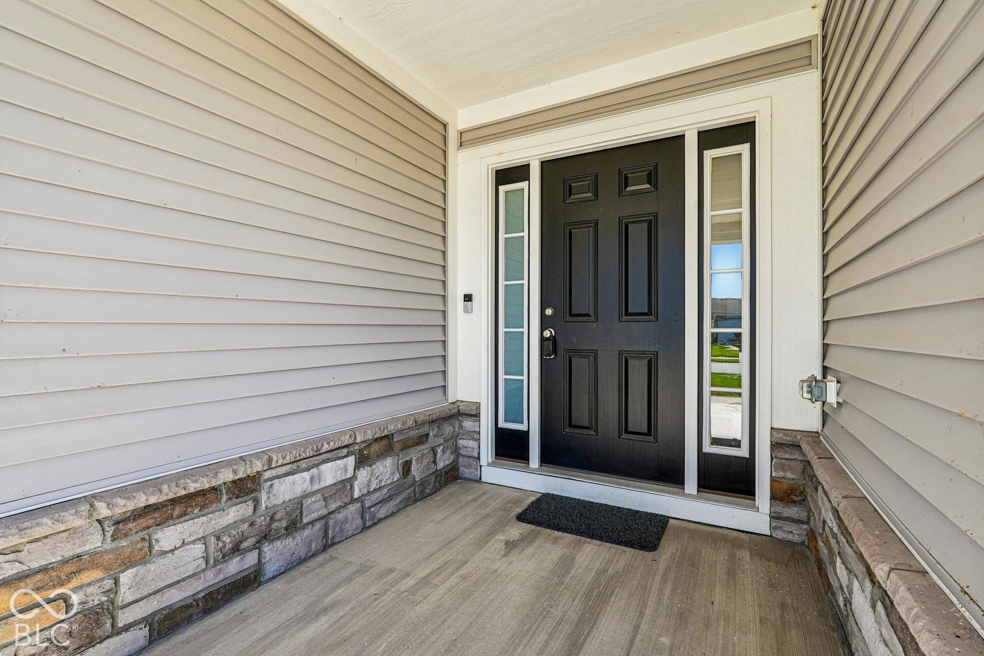 Covered front porch with concrete flooring, stone veneer accents, light-colored siding, and a paneled entry door with sidelights