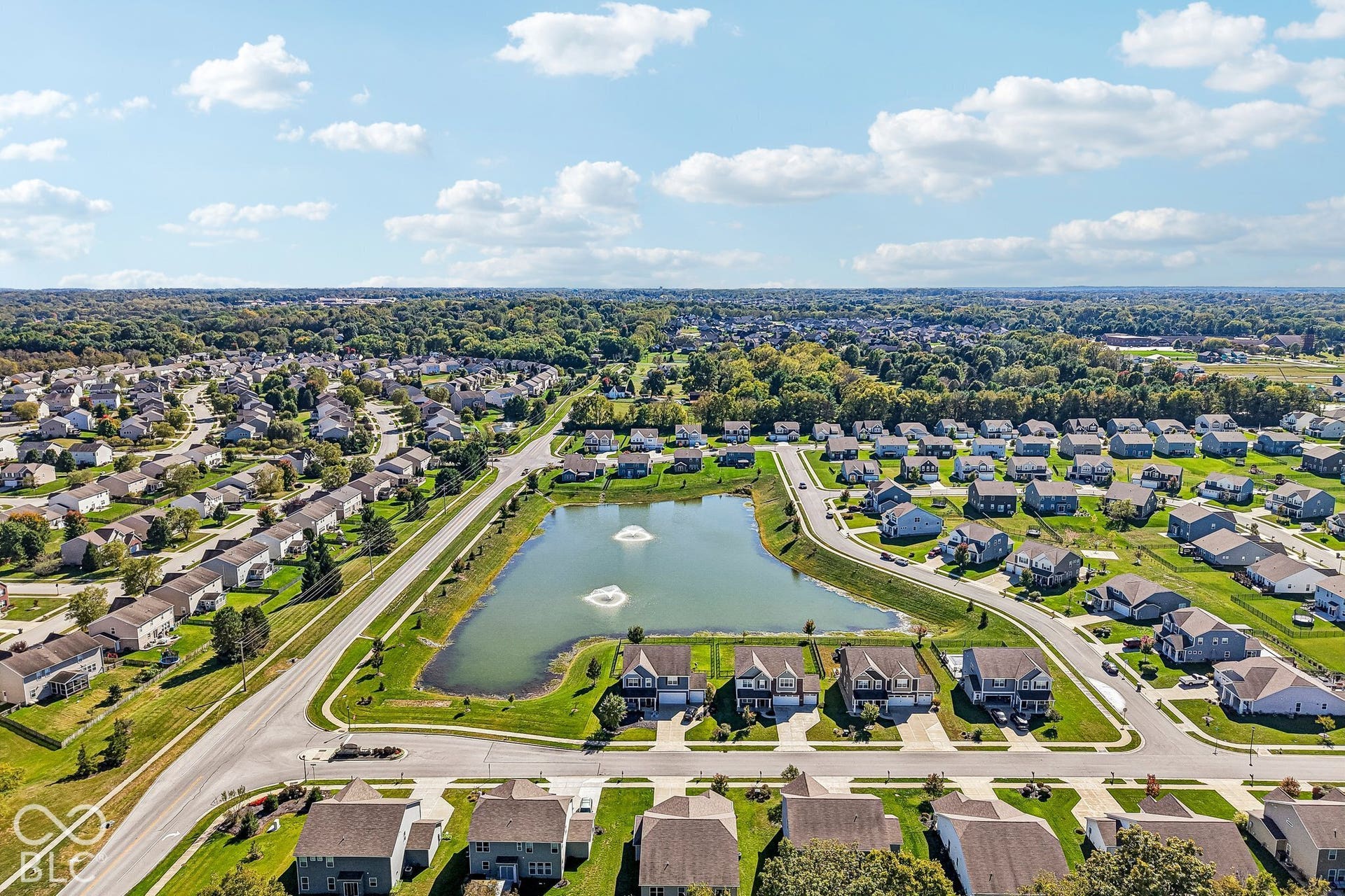 Expansive residential community featuring a large pond with fountains, paved roadways, and surrounding green spaces