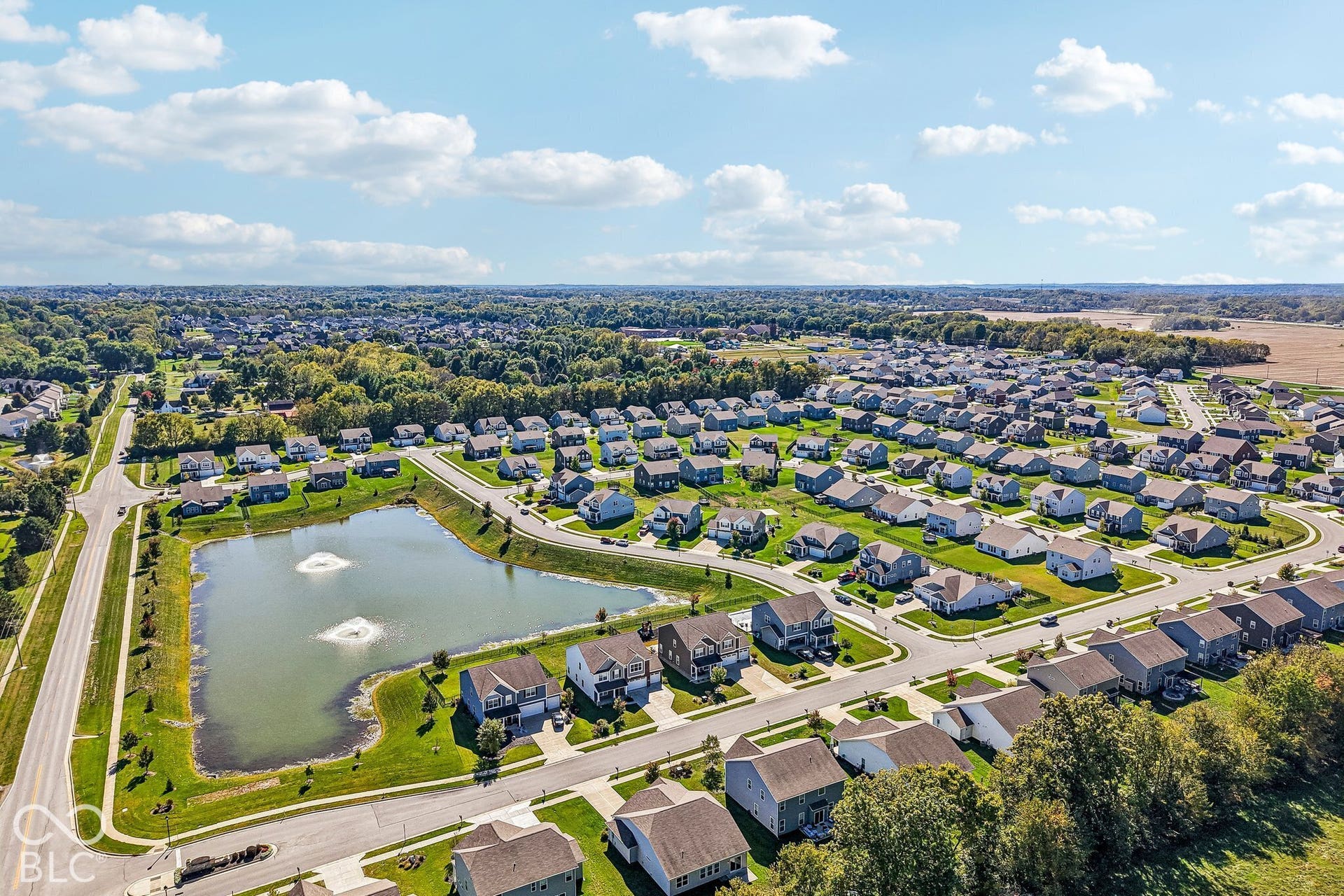 Residential development featuring a central pond with fountains, surrounded by paved roads and established homes, set against a backdrop of mature trees and open fields