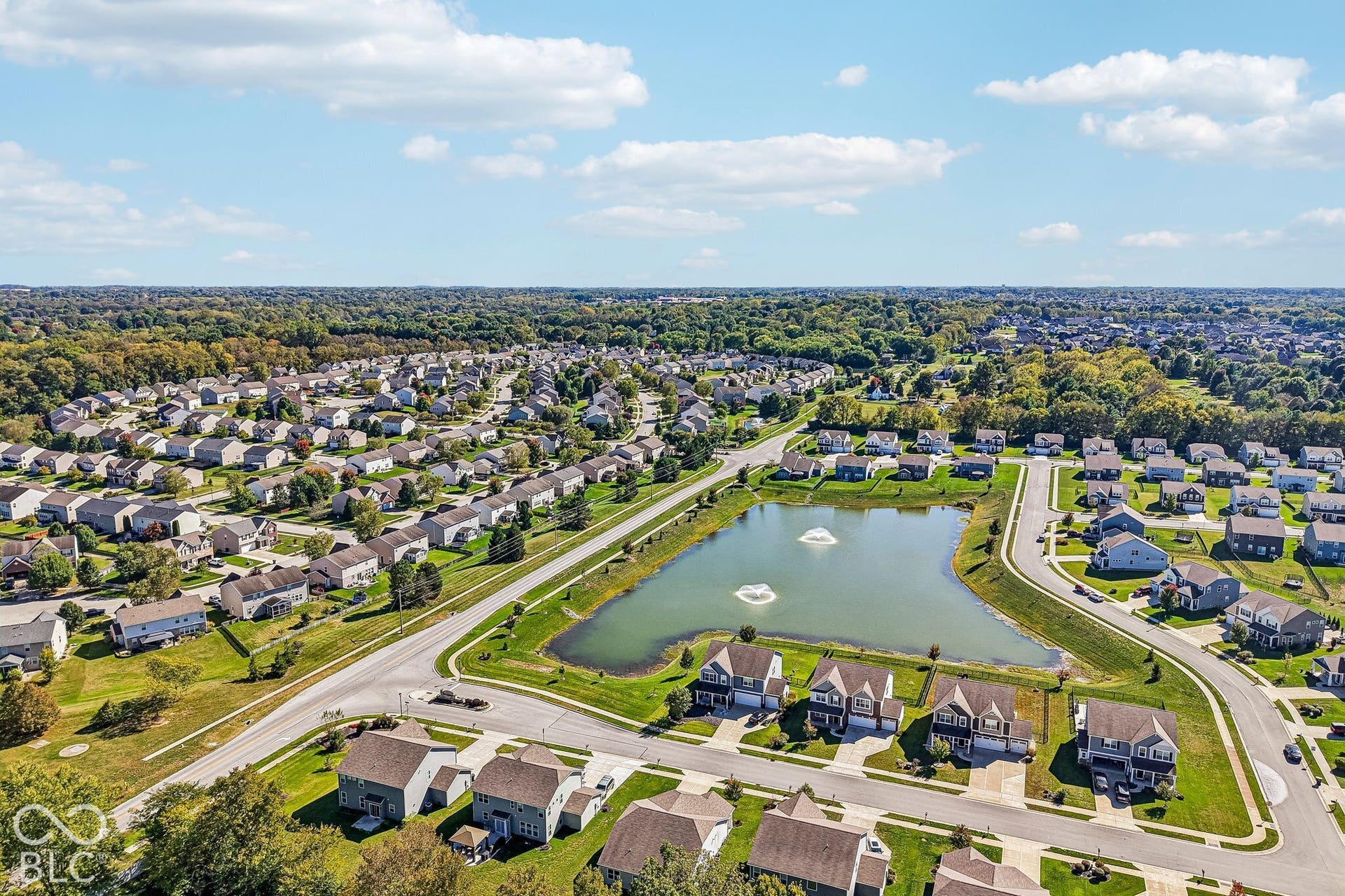 Residential neighborhood with uniform housing styles, featuring a central pond with two fountains, surrounded by mature trees