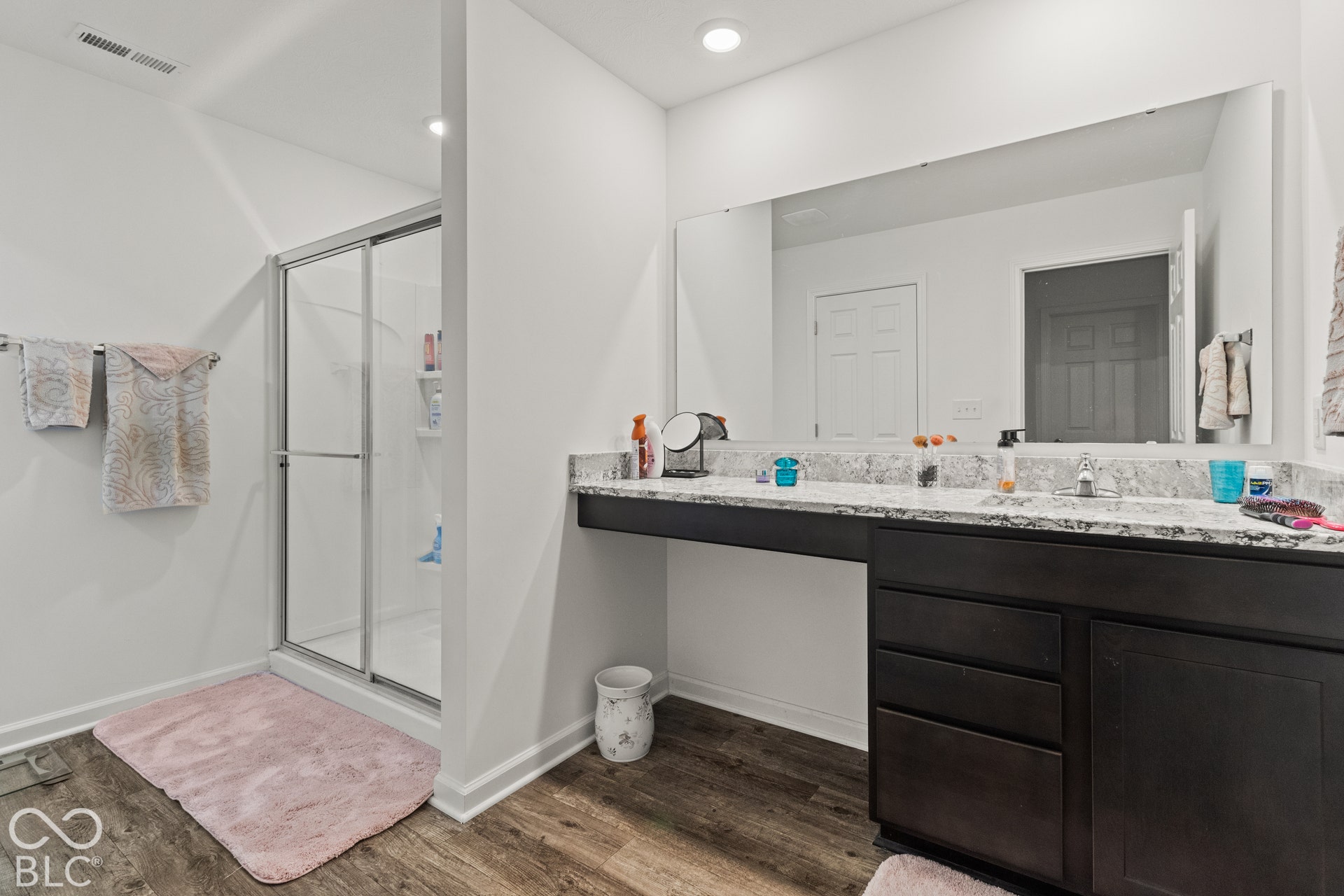 Wood-finish flooring, glass-enclosed shower, dark wood-finish vanity with drawer storage, light-toned countertop, and a large wall-mounted mirror