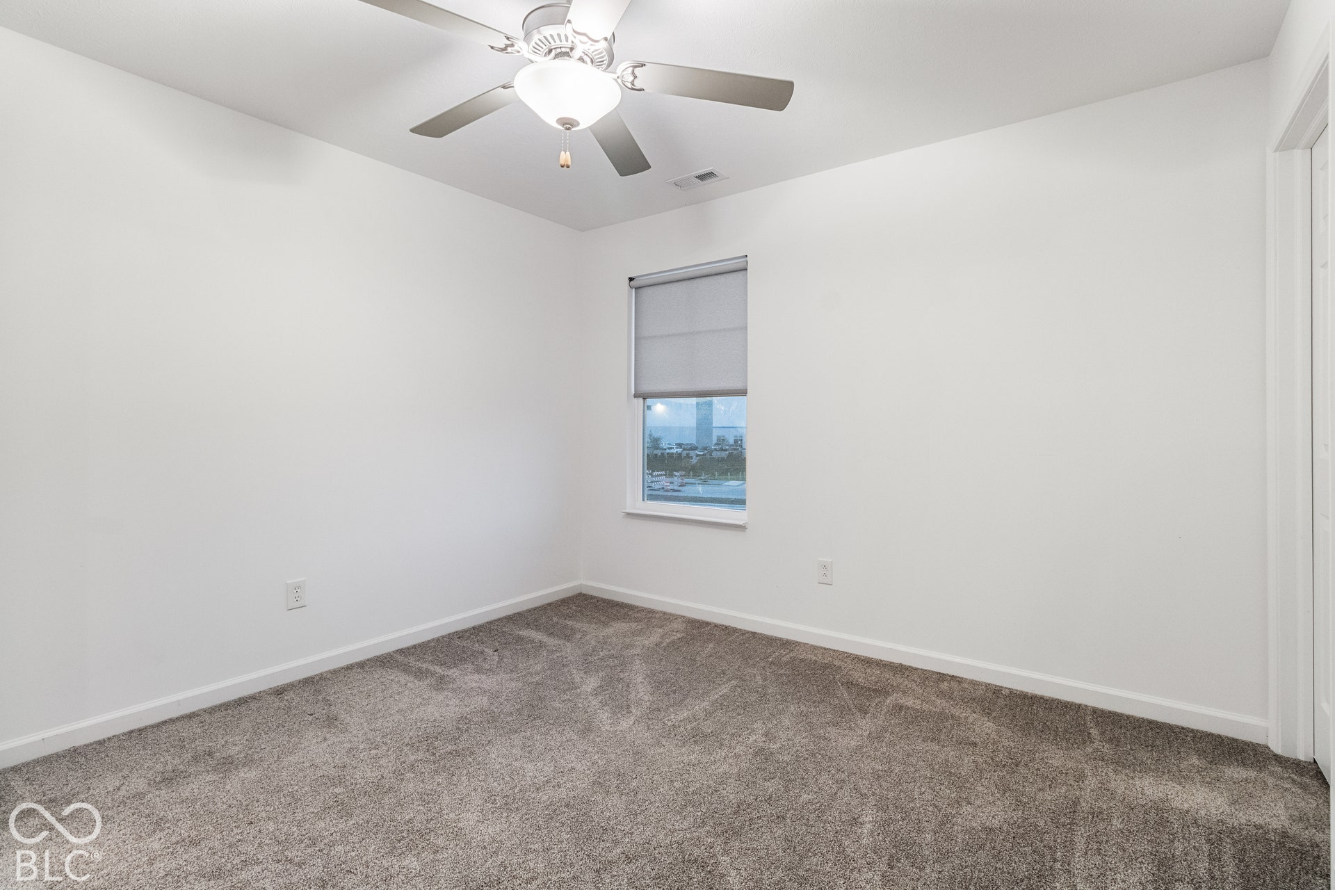 Neutral wall color, window with roller shade, gray carpet, and ceiling fan with light fixture