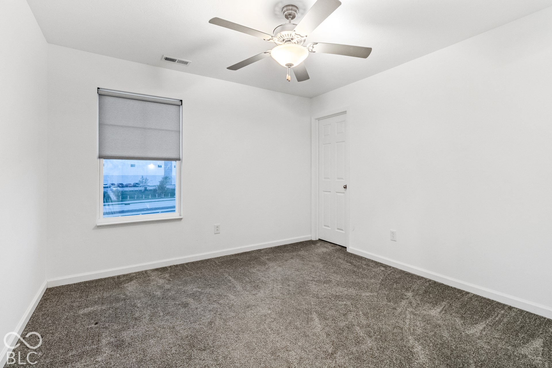 Gray carpeting, white walls, white trim, single window with a roller blind, ceiling fan with integrated lighting, and a white paneled door
