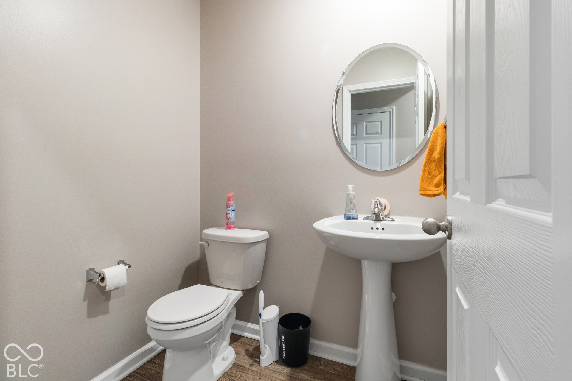 Pedestal sink, wall-mounted circular mirror, white toilet, and wood-finish flooring