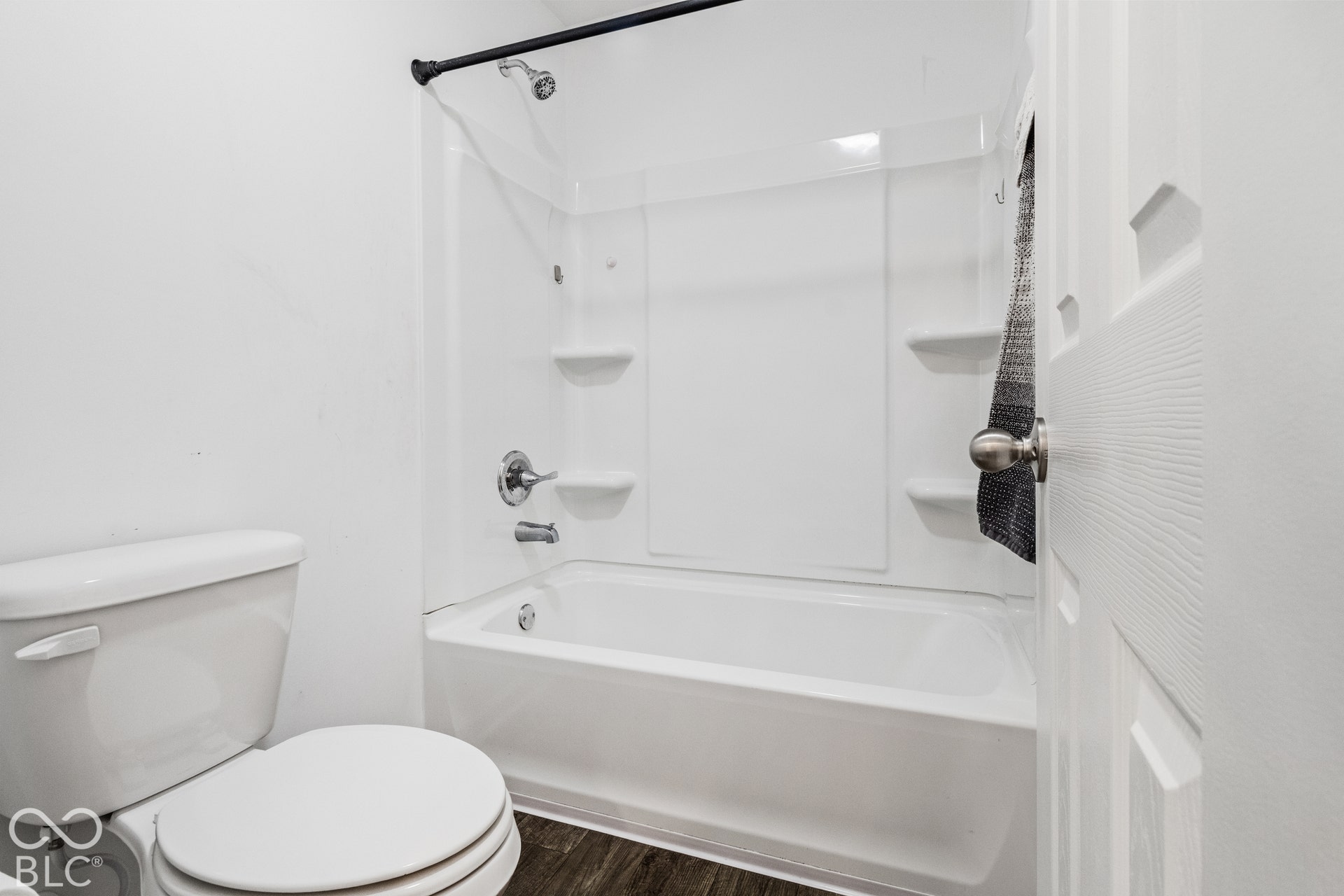 White bathtub and shower combination unit with integrated shelving, chrome fixtures, and a dark wood-finish floor
