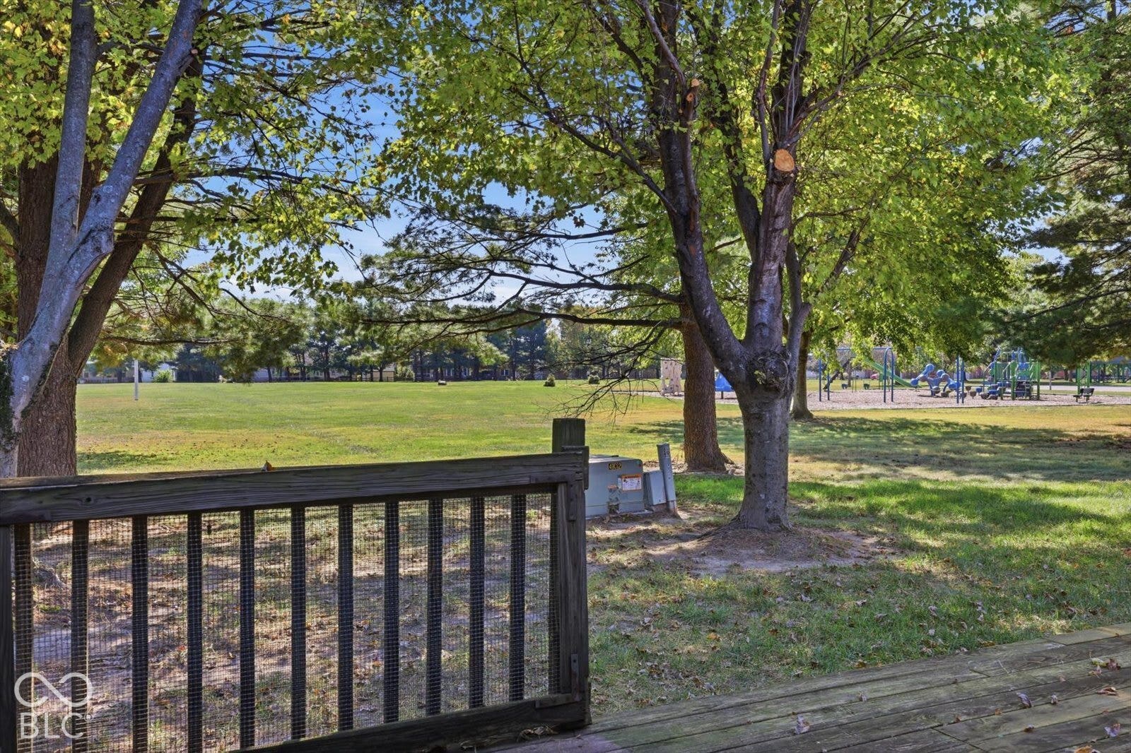 Wood-plank deck featuring a metal-railing enclosure, mature trees, and an open grass field