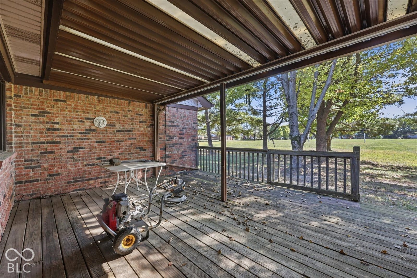 Covered wooden deck featuring a brick exterior wall, corrugated metal roofing, and a wooden railing