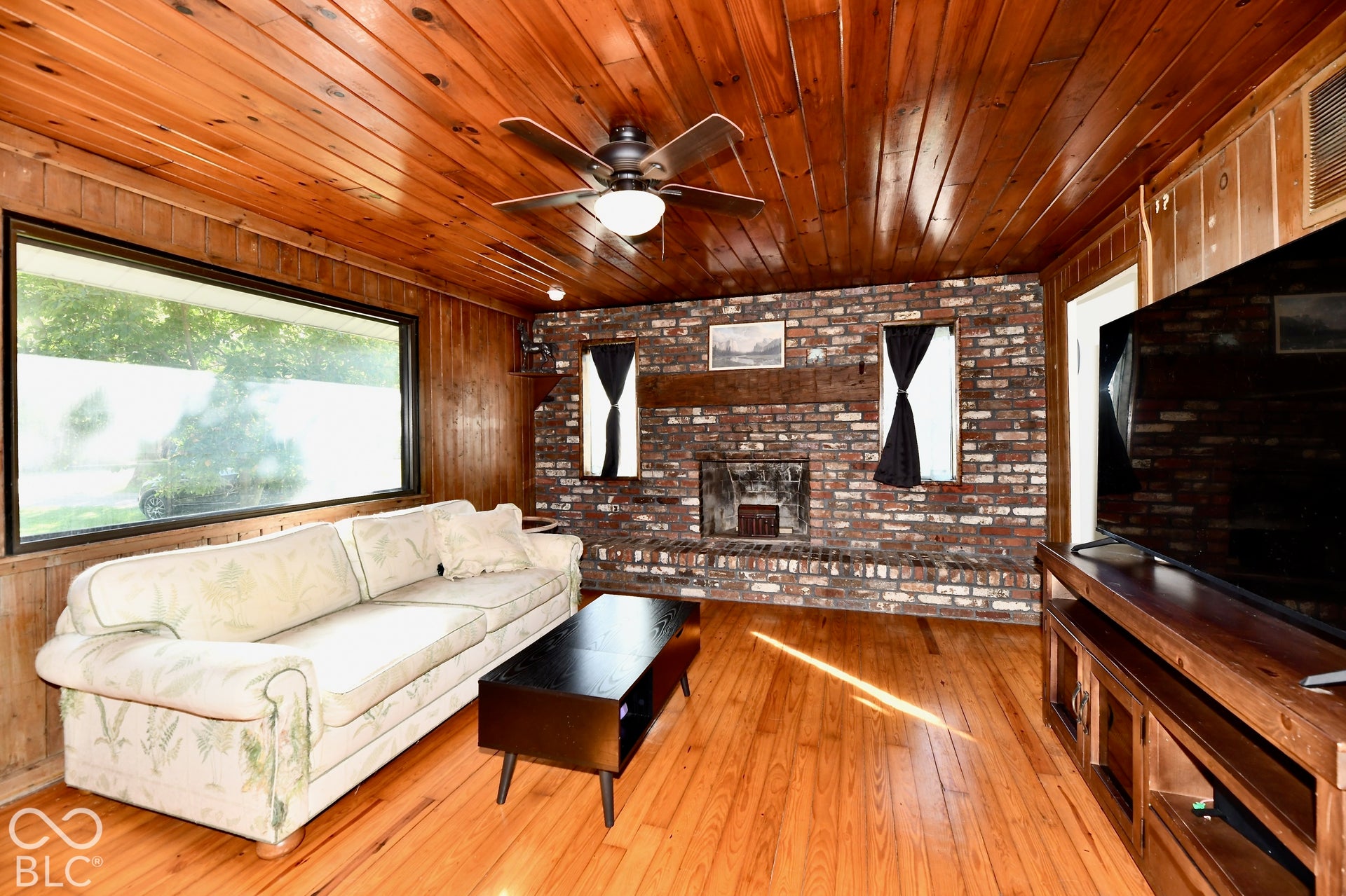 Wood-paneled walls and ceiling, brick-faced fireplace with a wood mantle, wood-finish flooring, and a ceiling fan with integrated lighting