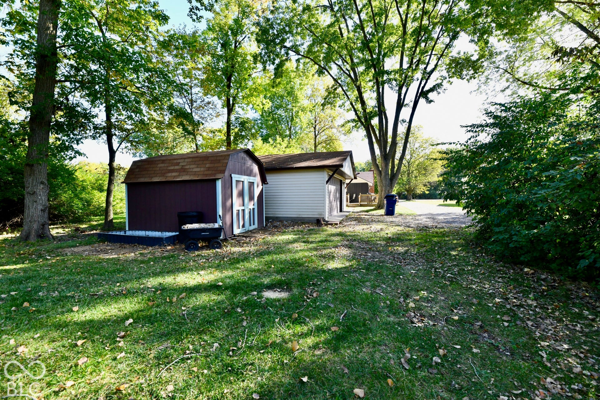Two detached storage sheds with shingle roofs, surrounded by mature trees and a grassy area