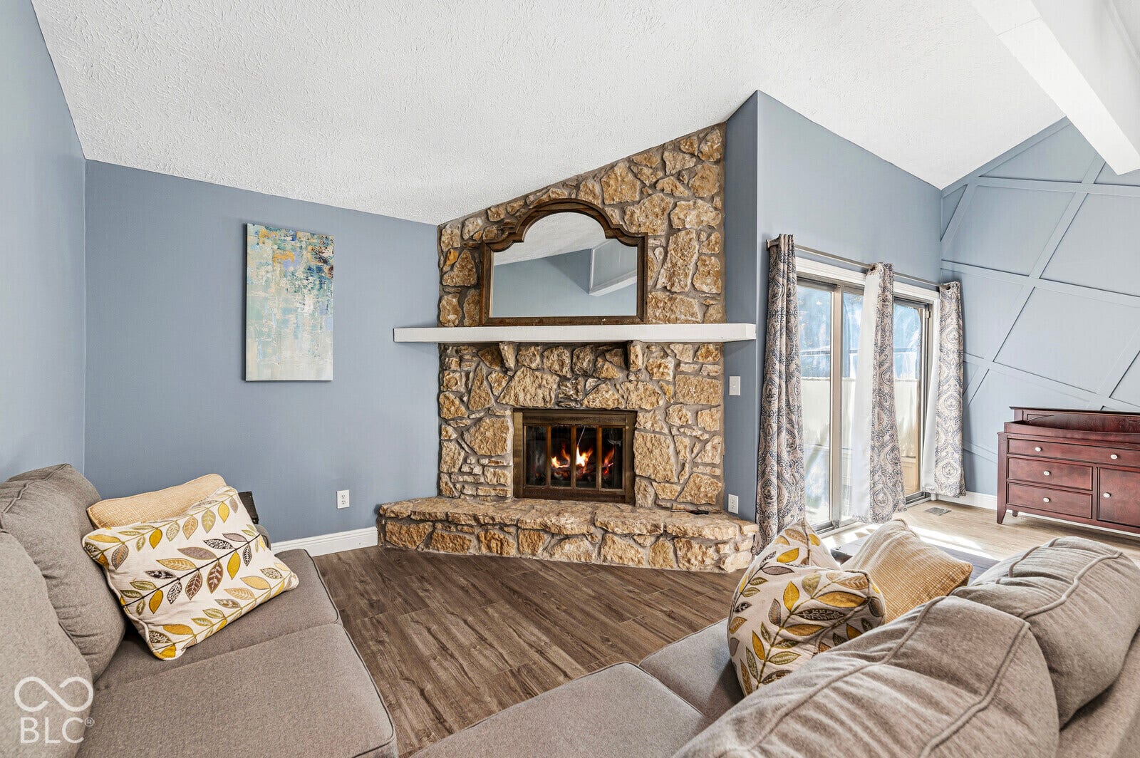 Spacious living area featuring a wood-finish plank floor, stone fireplace with built-in shelving and mantel, vaulted ceiling with skylight, and glass sliding doors providing natural light