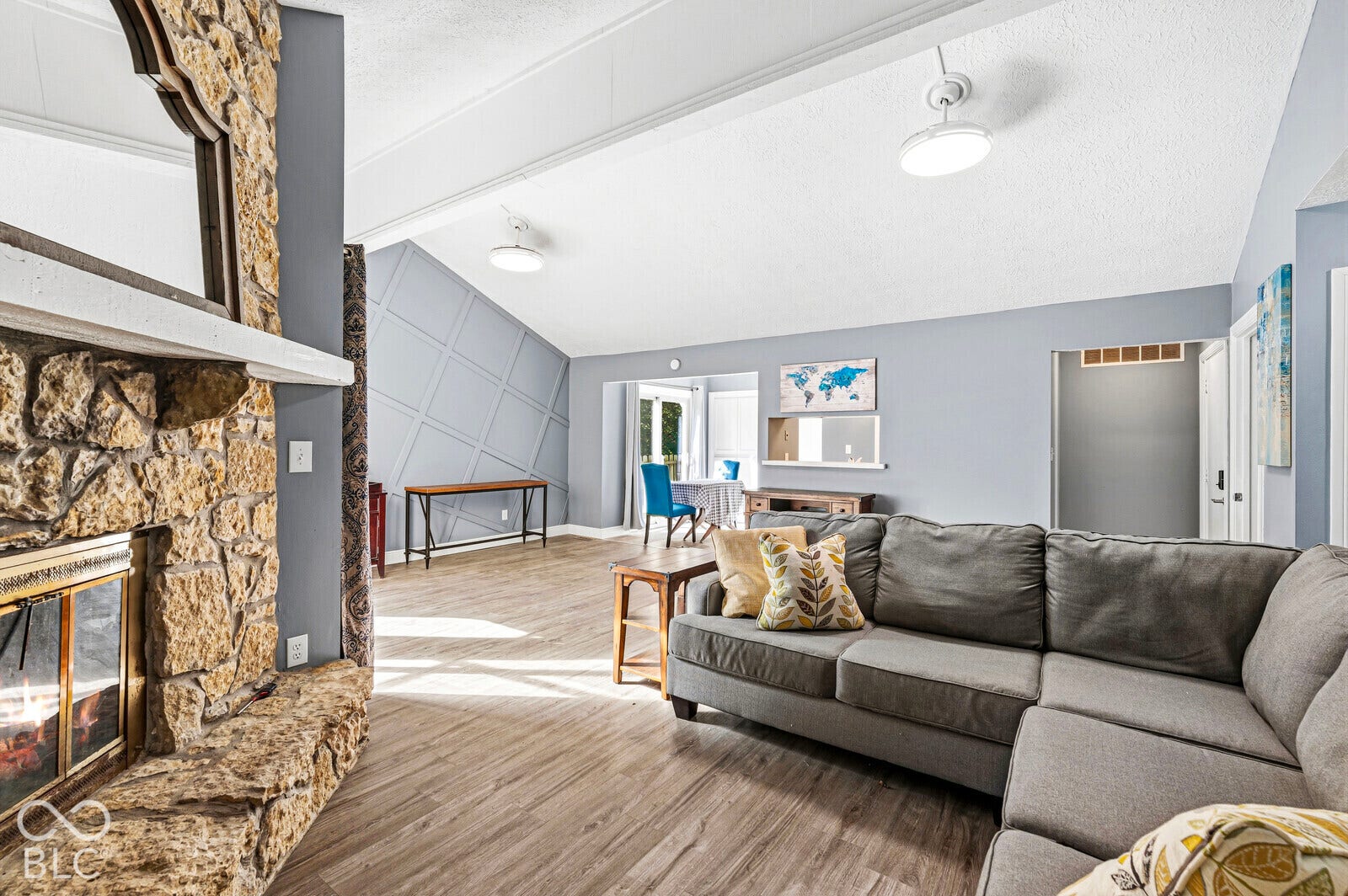 Spacious living area featuring a stone-clad fireplace with a hearth, vaulted ceilings, wood-finish flooring, and a decorative accent wall