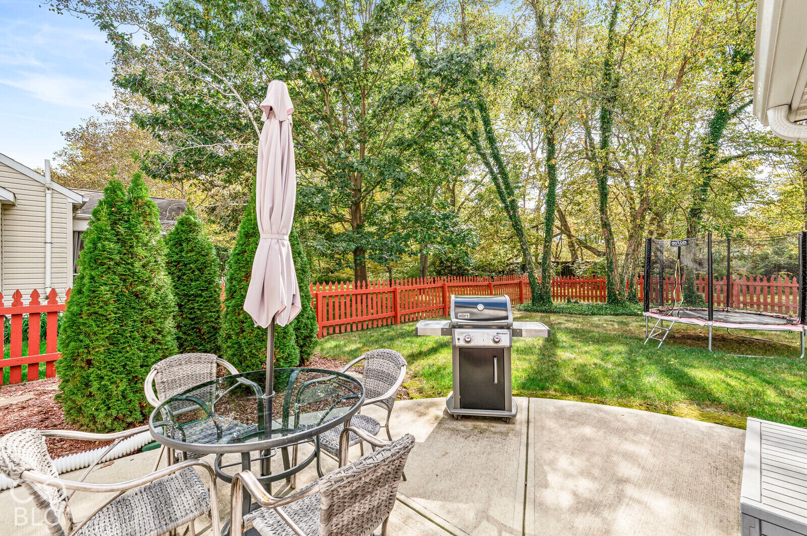 Concrete patio featuring a perimeter red picket fence, mature trees, and landscaping
