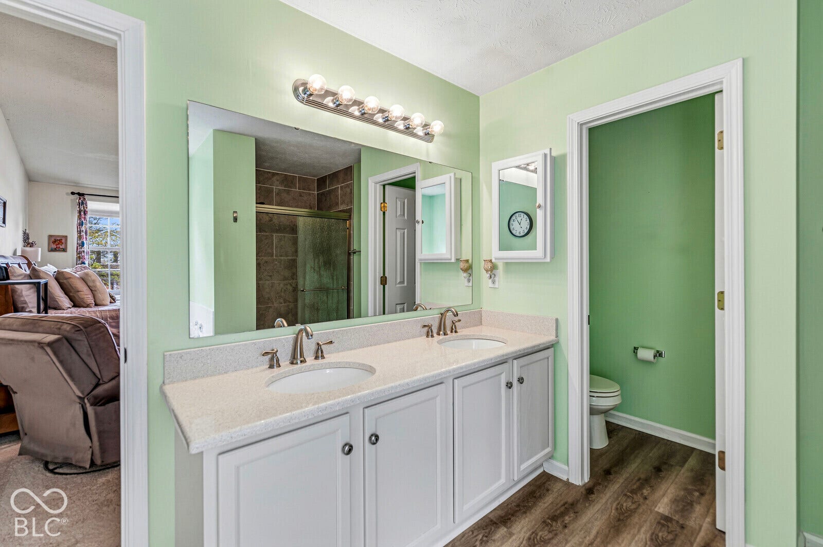 Dual-sink vanity featuring a light-toned solid surface countertop, white shaker-style cabinetry, a wall-to-wall mirror, six-light fixture, and wood-finish flooring