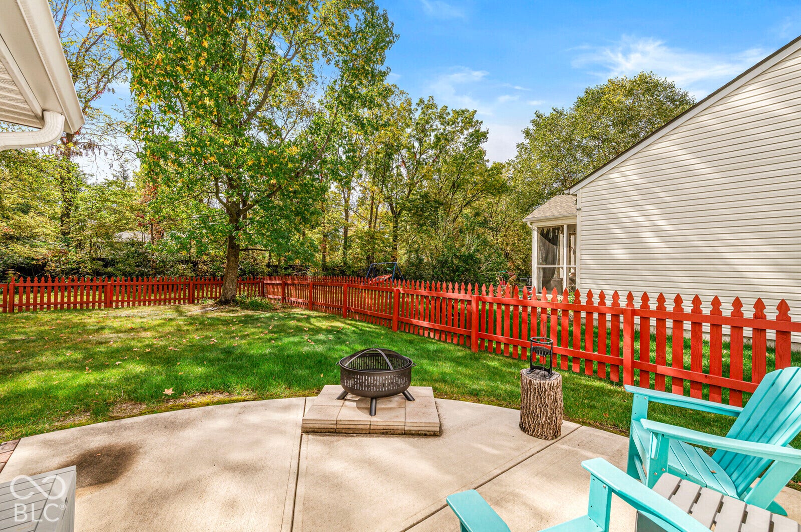 Backyard featuring a concrete patio, stone fire pit base, wood picket fencing, and mature trees