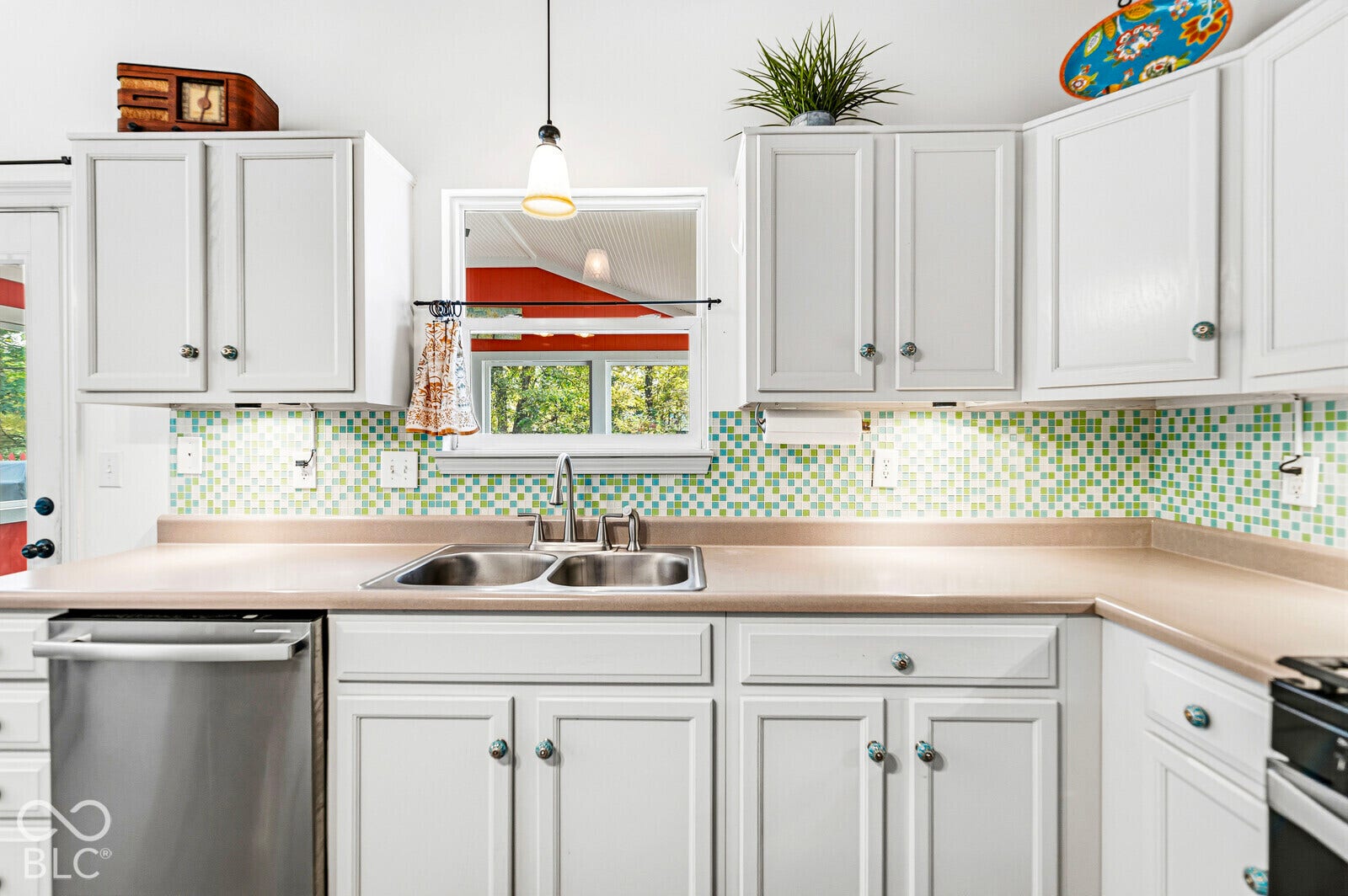 Kitchen featuring white shaker-style cabinetry, a multi-colored mosaic tile backsplash, solid-surface countertops, a stainless steel double-basin sink, and a pendant light fixture