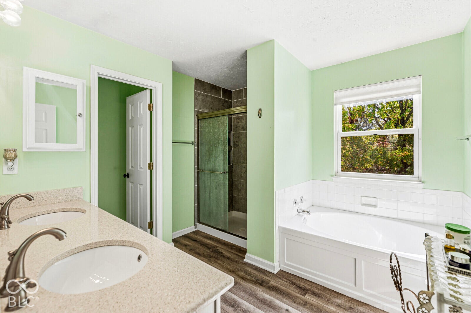 Dual-sink vanity with a solid-surface countertop, walk-in shower with tiled surround, and a deep soaking tub featuring white tile accents and wood-finish flooring