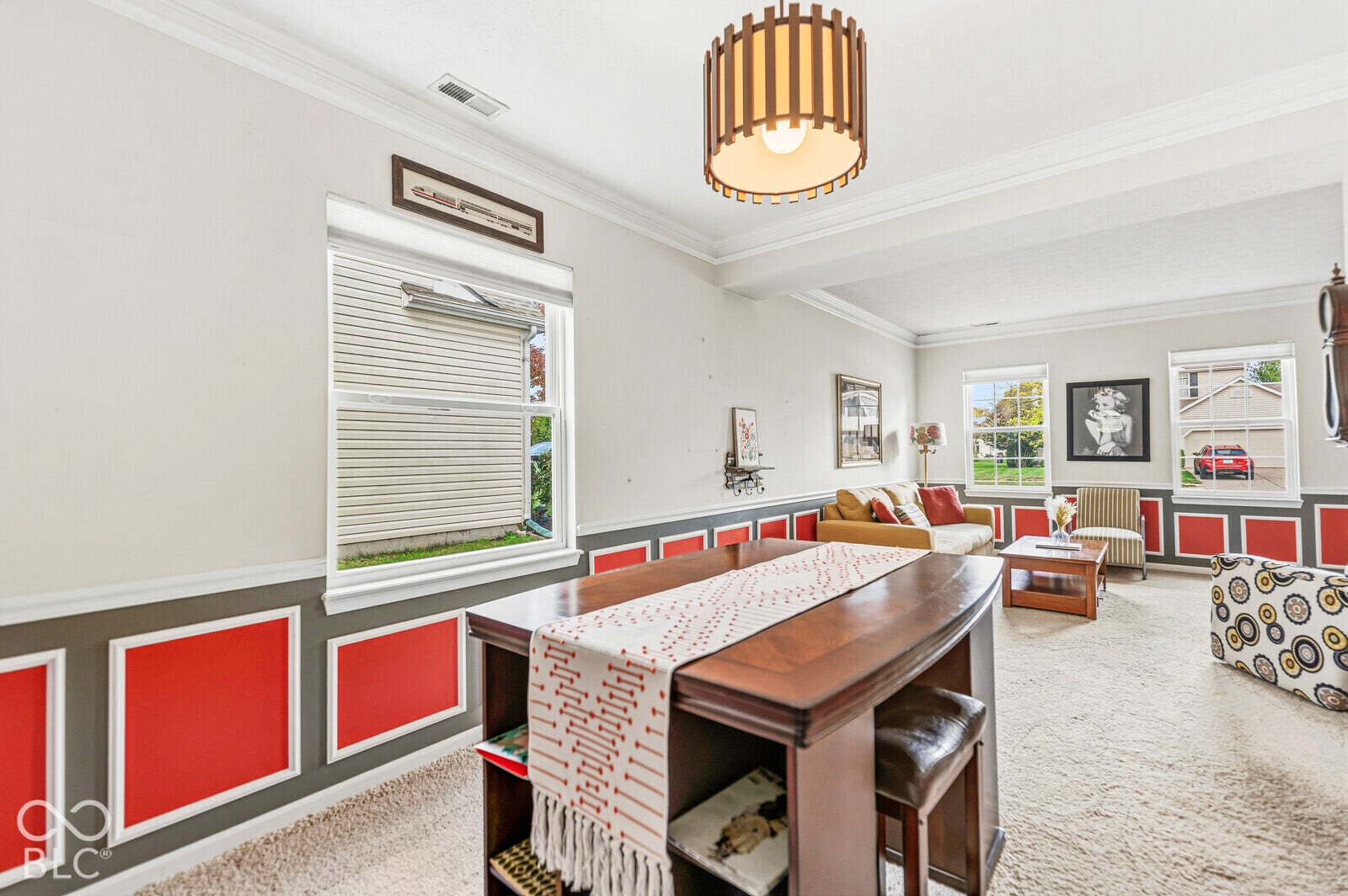 Living area featuring neutral carpet, decorative wainscoting with paneled detailing, crown molding, and a cylindrical slatted pendant light fixture