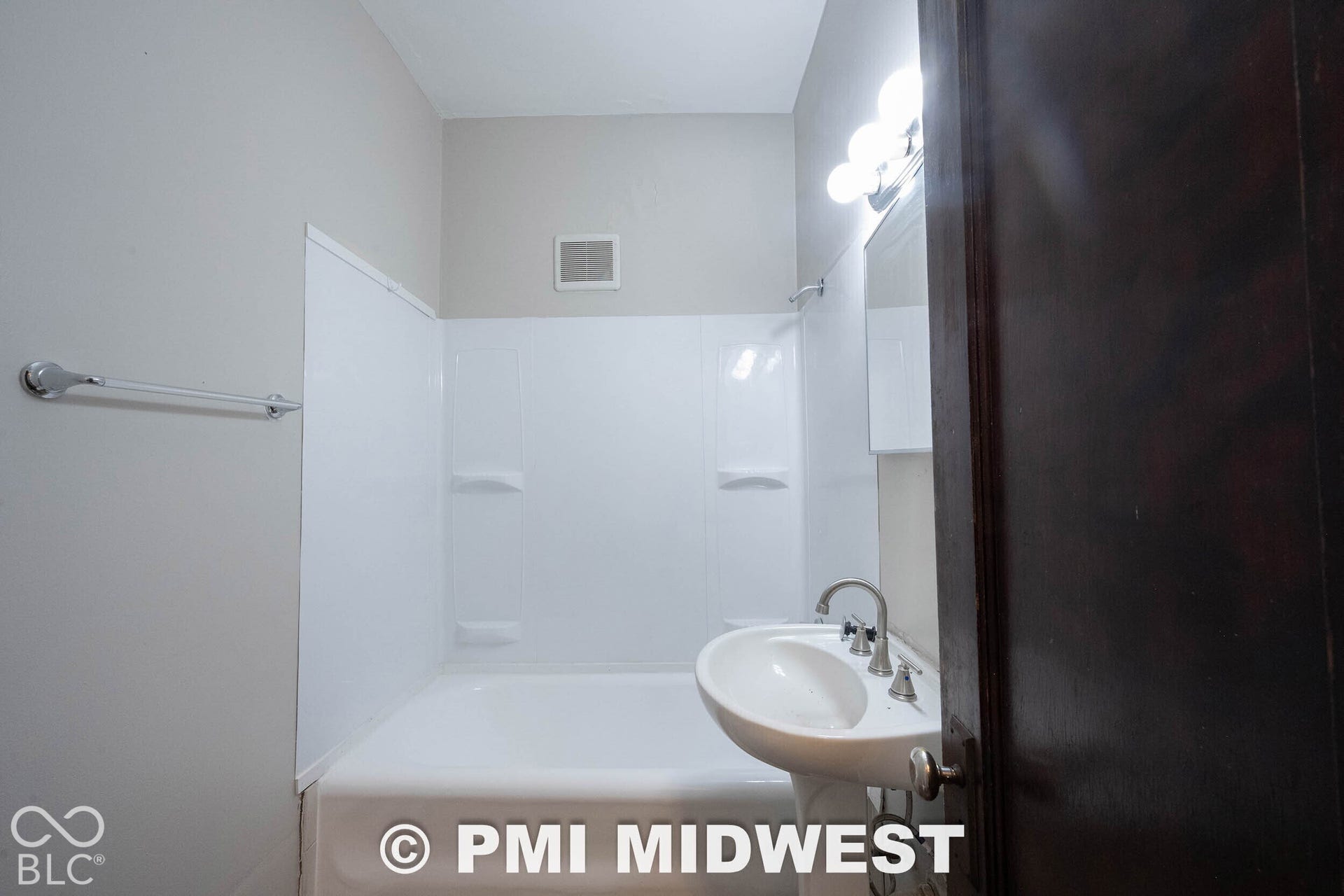 Pedestal sink with chrome fixtures, white tub with shower surround, and vanity lighting