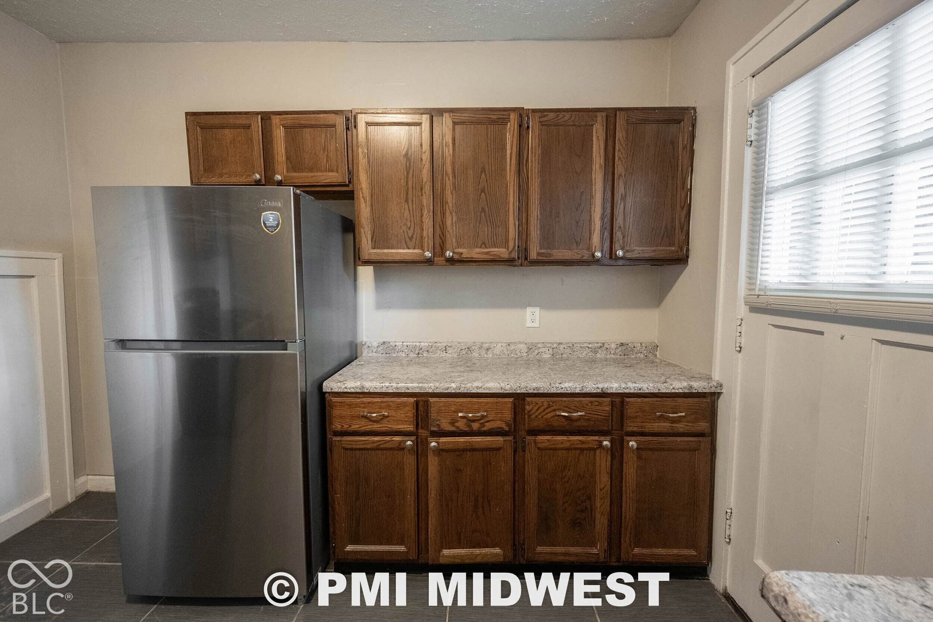 Stainless steel refrigerator, wood-finish cabinetry, light-toned countertop, and dark tile flooring