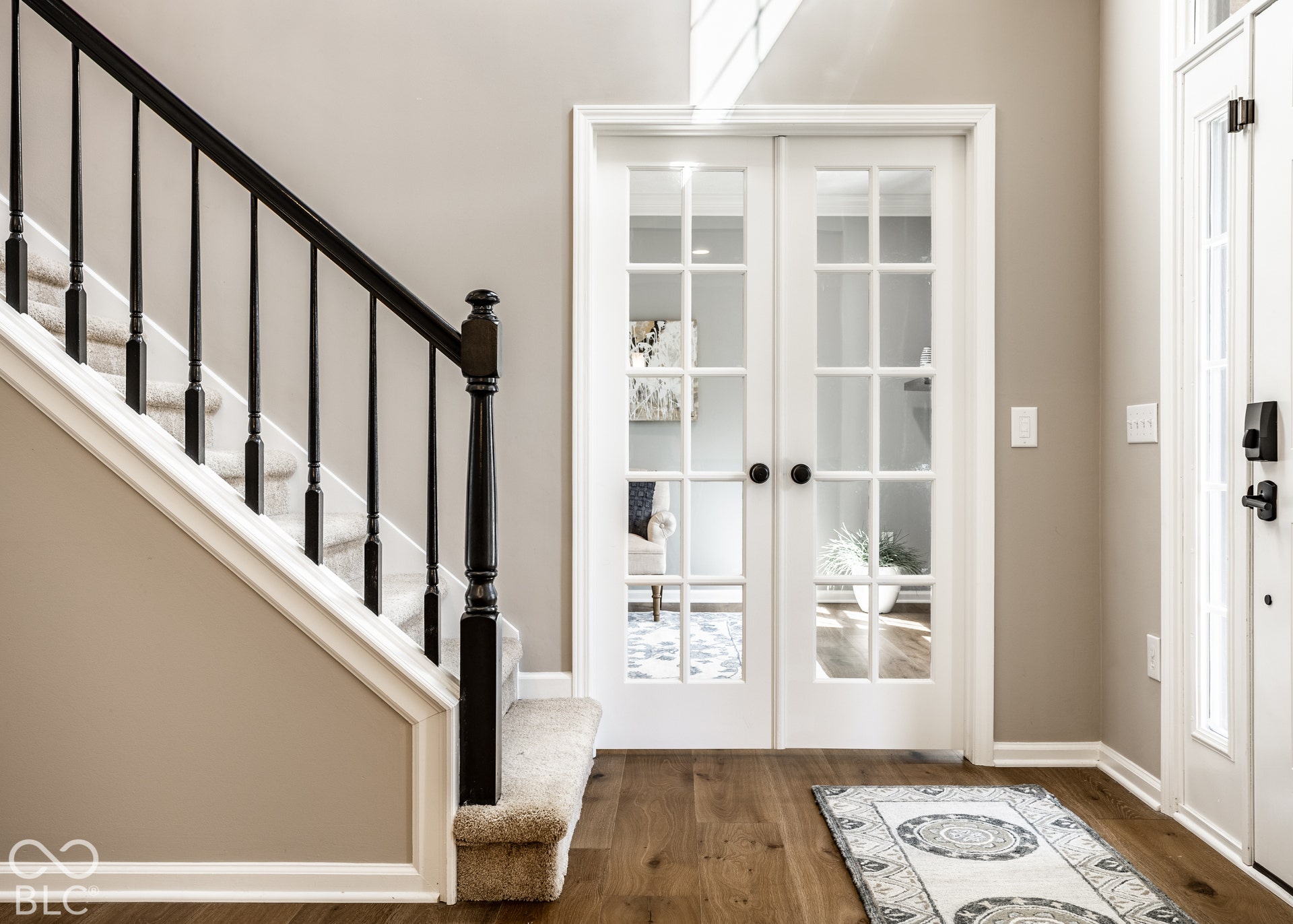 Wood-finish flooring with white baseboards, a carpeted staircase with dark wood balusters, and white French doors with dark hardware