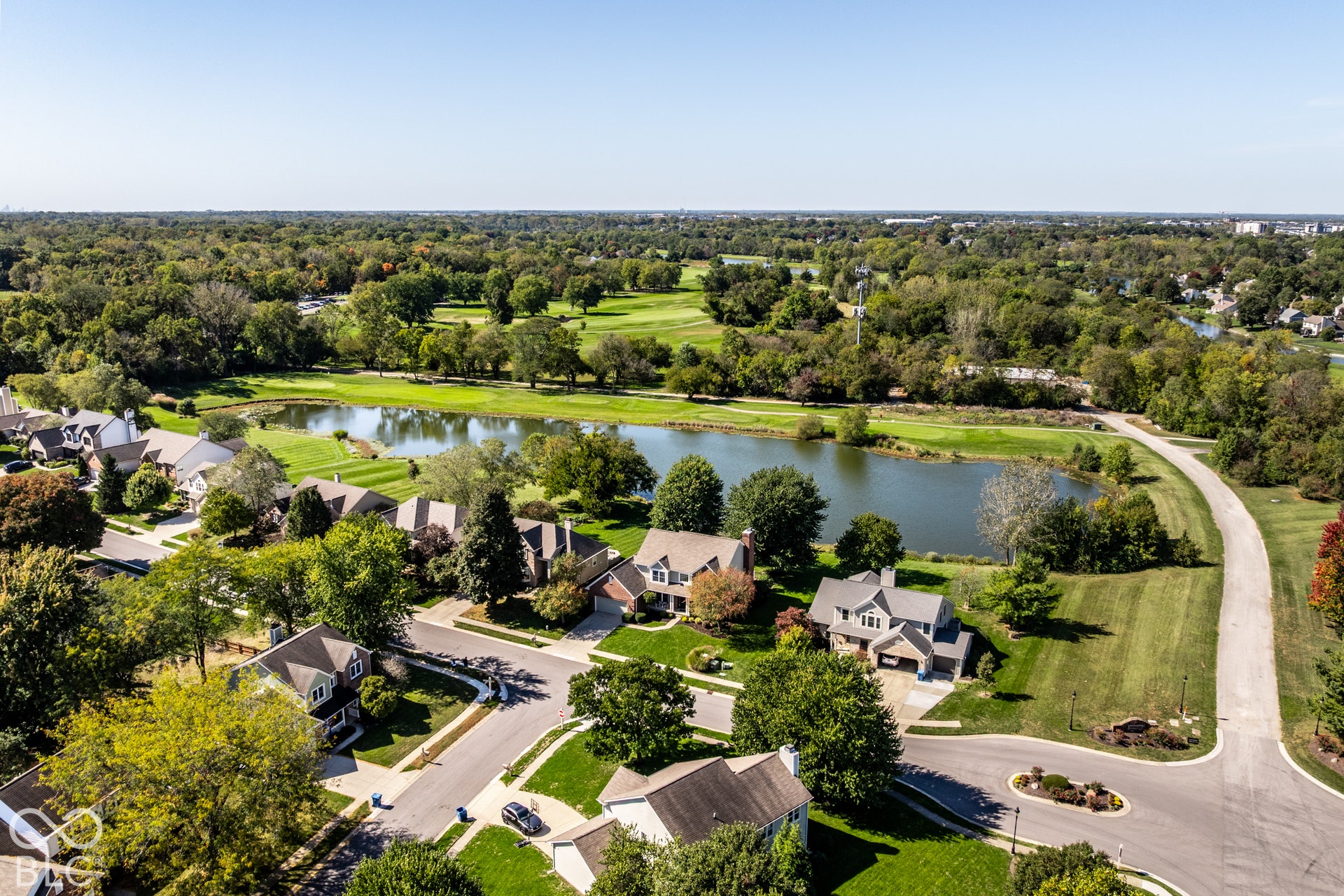 Aerial neighborhood perspective featuring a central pond, surrounding residential properties, extensive green spaces, and a distant golf course