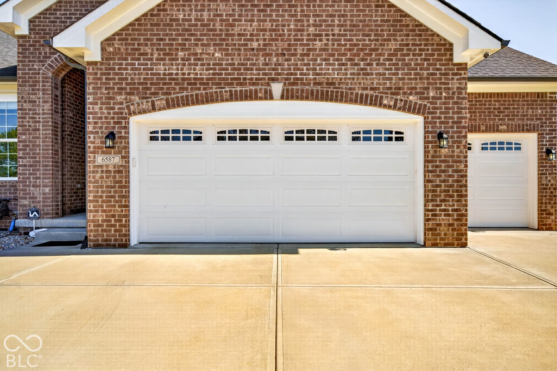 Brick exterior with white garage doors, featuring upper window panels, decorative exterior lighting, and a concrete driveway