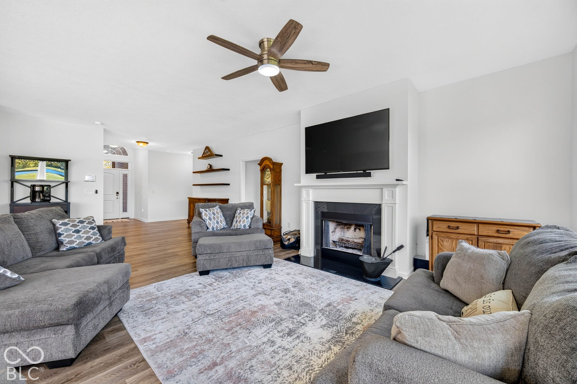 Living area featuring wood-finish flooring, a ceiling fan, a fireplace with a white mantel, and a media wall
