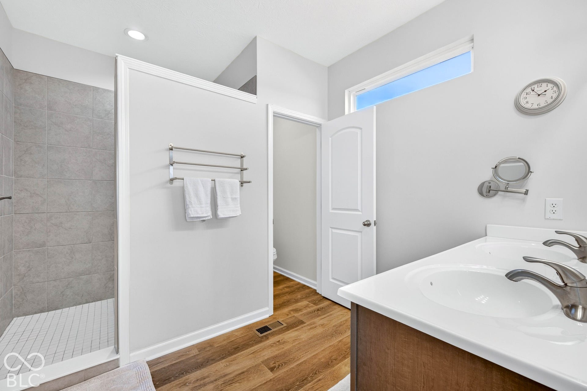 Walk-in shower with gray tile surround, wood-finish flooring, dual vanity with white countertops, and a transom window