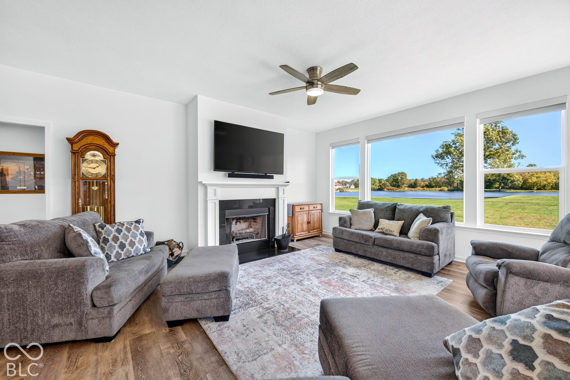 Spacious living area featuring a central fireplace with a mantel, wood-finish flooring, a ceiling fan with integrated lighting, and expansive windows showcasing a prominent waterfront