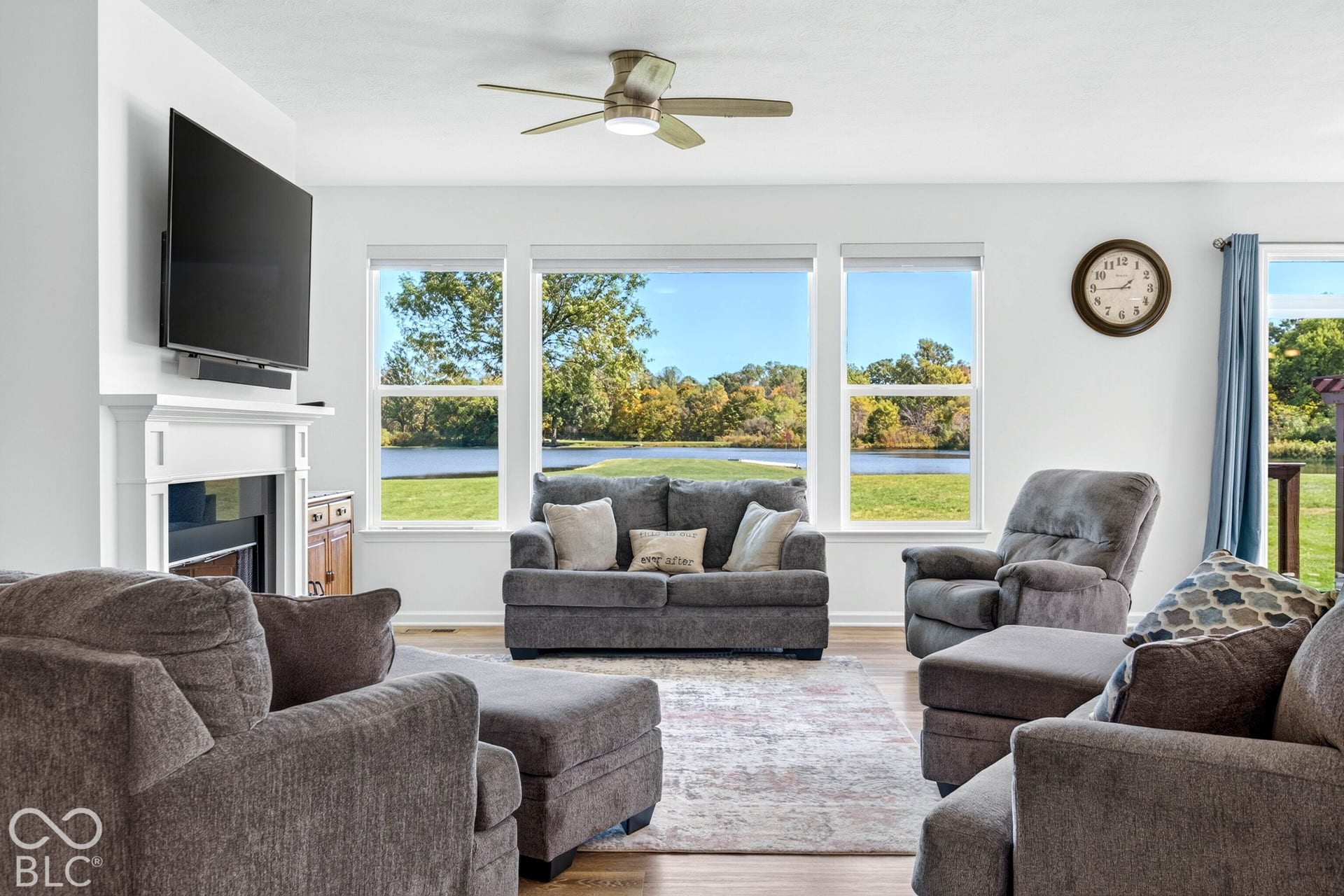 Living area featuring a white mantel, ceiling fan, and large windows with water and treeline scenery