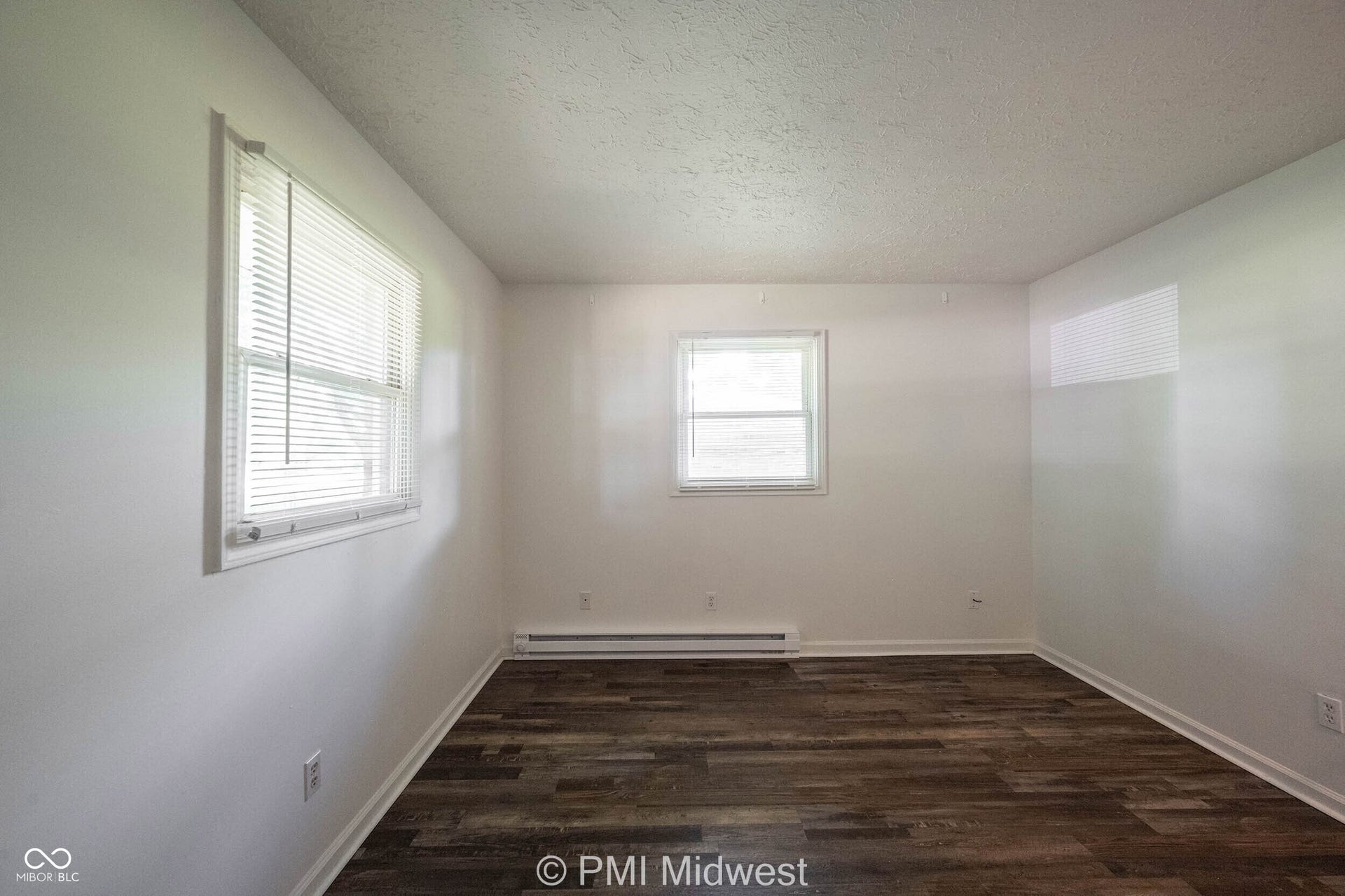 Spacious room featuring wood-finish flooring, white walls, two windows with blinds, and baseboard heating