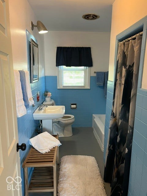 Pedestal sink, wall-mounted mirror, ceramic tile wainscoting, built-in bathtub, and window with blinds