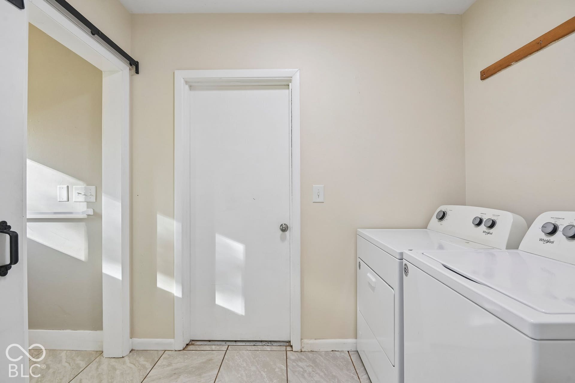 Dedicated laundry room featuring side-by-side washer and dryer units, tile flooring, abundant natural light from a transom window, and direct access via a barn-style sliding door with black hardware