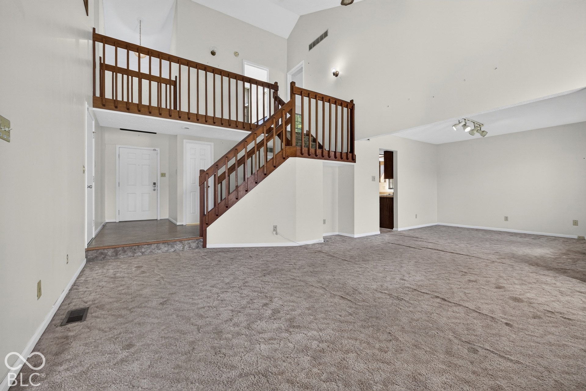 Vaulted ceilings with track lighting, an open-concept living area with carpet flooring, and a two-story entryway featuring a wood-finish staircase and balcony