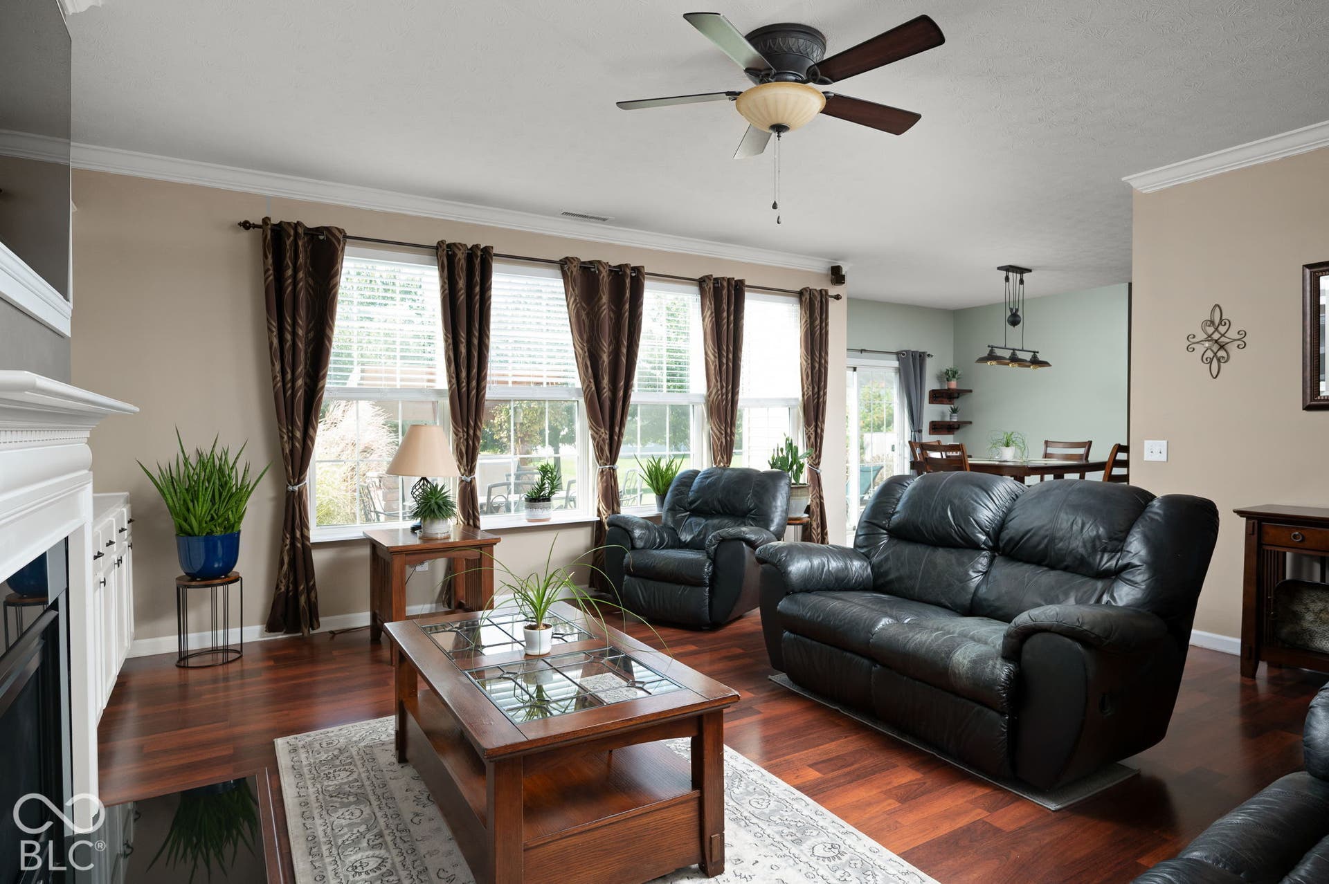 Bright, light-filled living room featuring expansive multi-pane windows with brown drapery, wood-finish flooring throughout, a ceiling fan with integrated lighting, and a fireplace with white surround and mantel