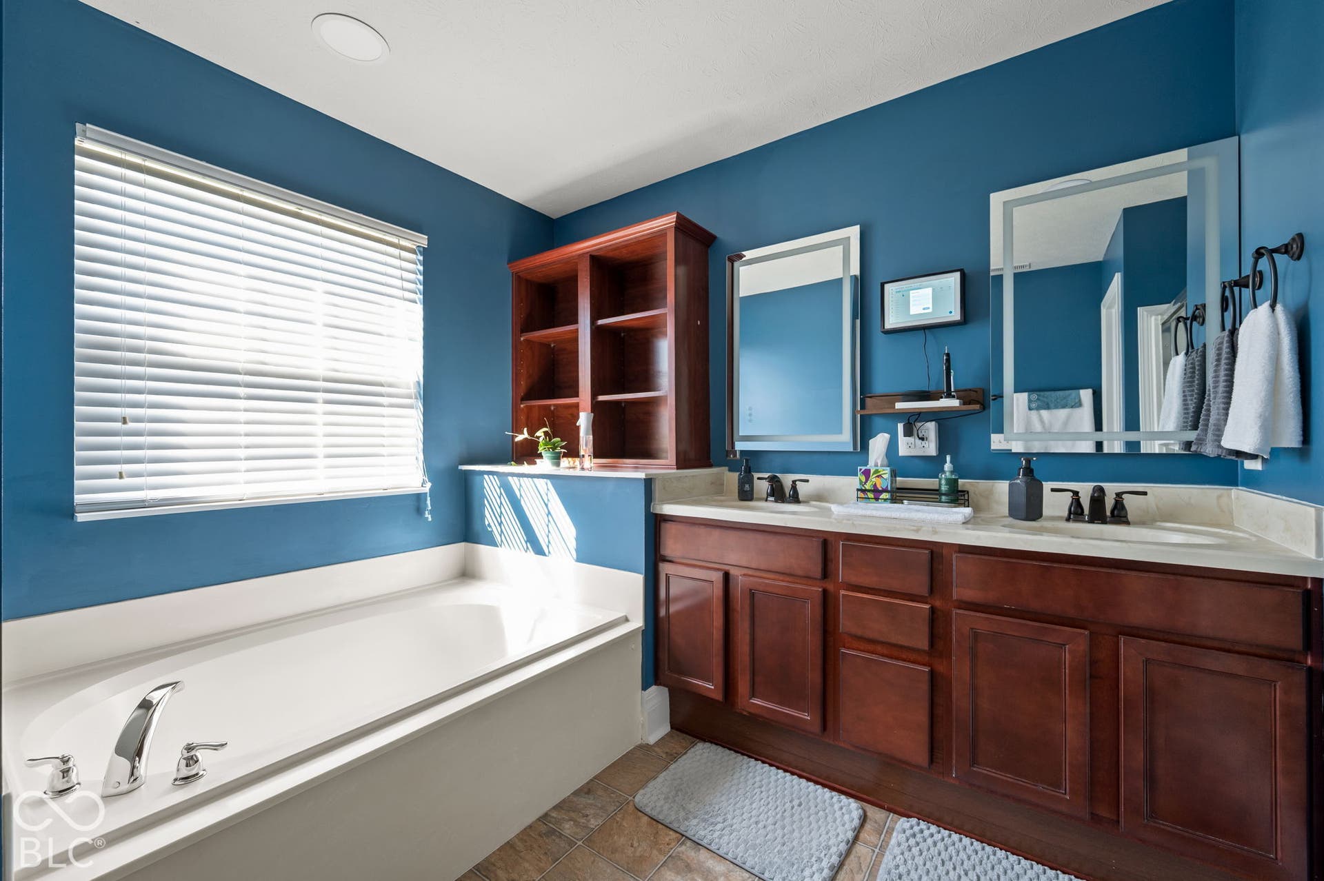 Dual vanity with integrated sinks, built-in shelving, a soaking tub, and a window with blinds