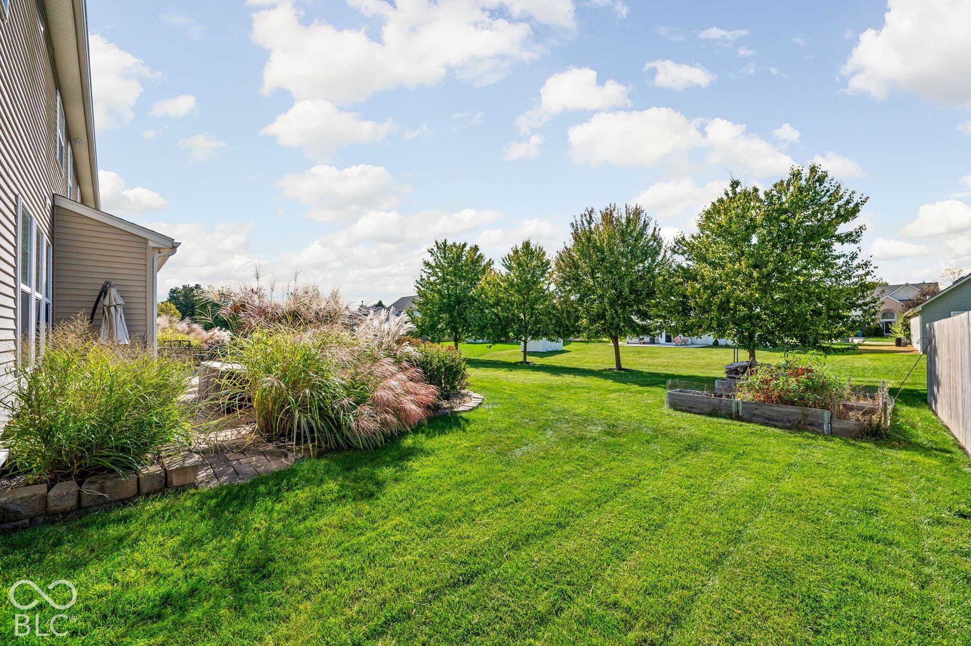 Expansive backyard featuring a stone patio, vibrant landscaping, green lawn, mature trees, and raised garden beds