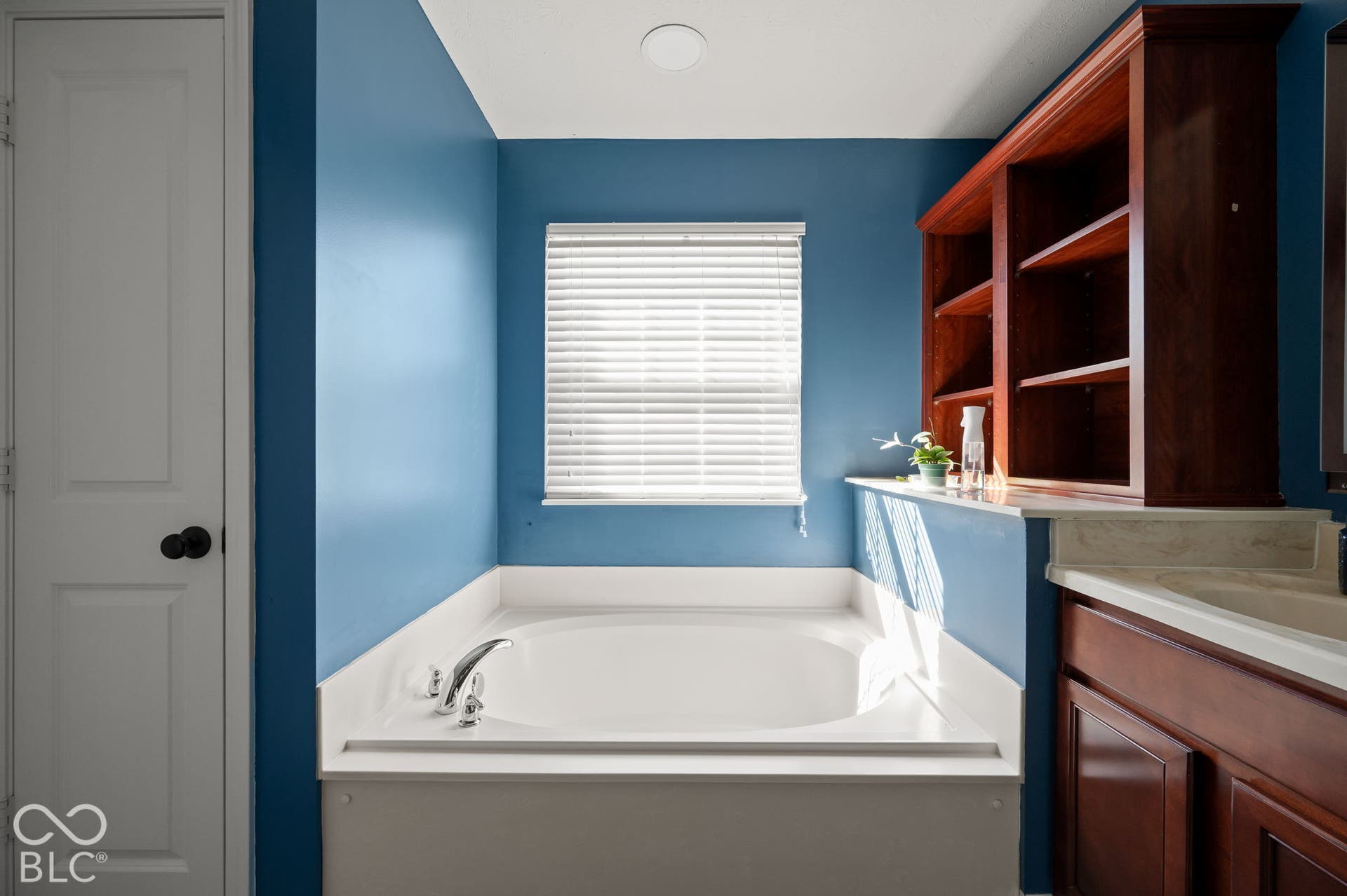 Built-in soaking tub with chrome fixtures, window with horizontal blinds, and a built-in wood-finish shelving unit