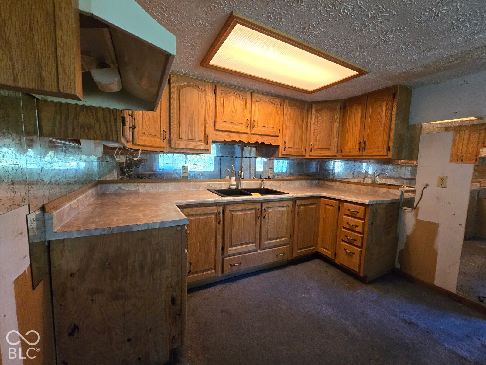 L-shaped countertops with a built-in sink, wood-finish cabinetry, overhead fluorescent lighting, and mirrored backsplash