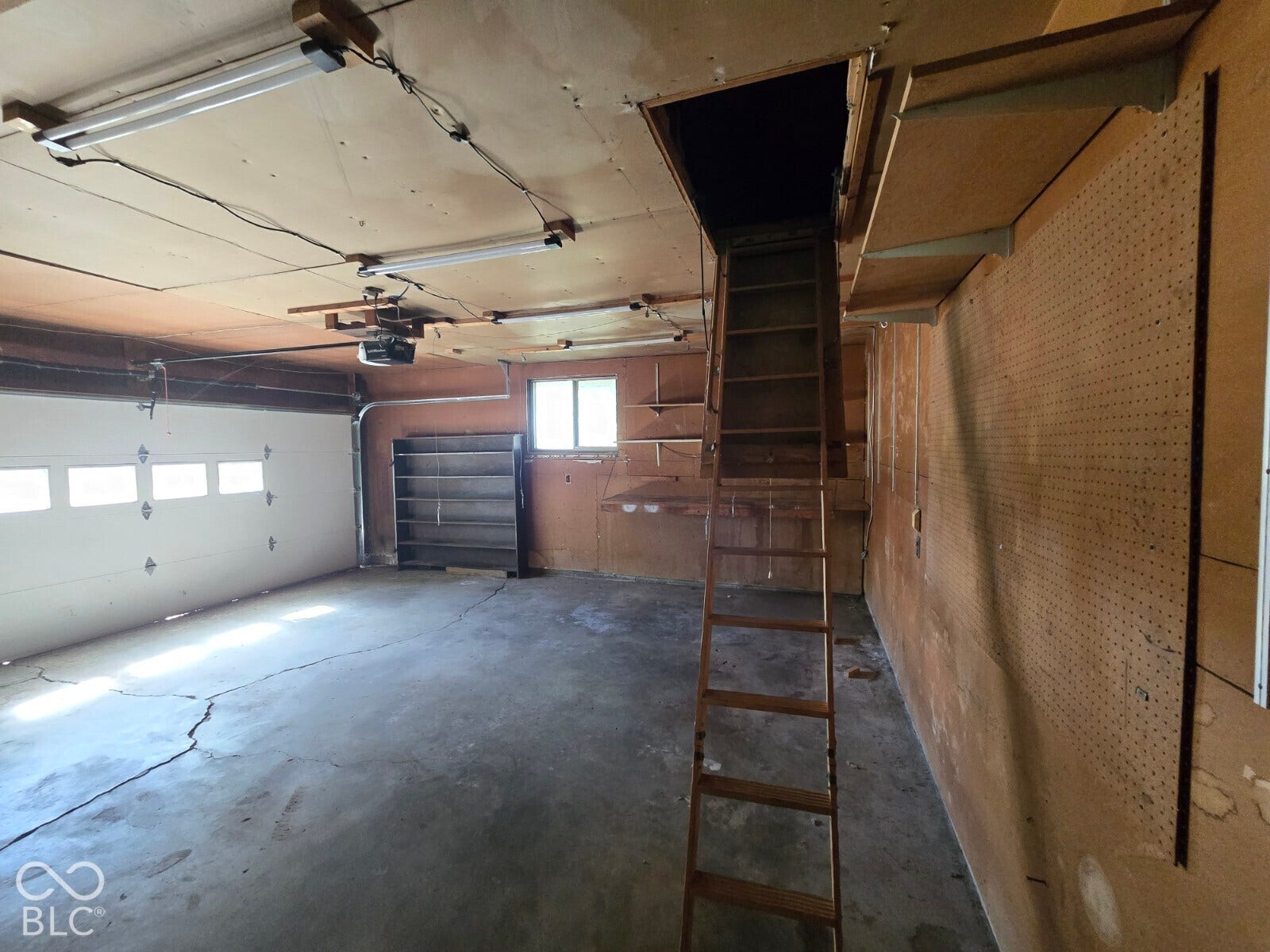 Attached garage space with a roll-up door, overhead fluorescent lighting, a built-in attic ladder, and pegboard wall organization